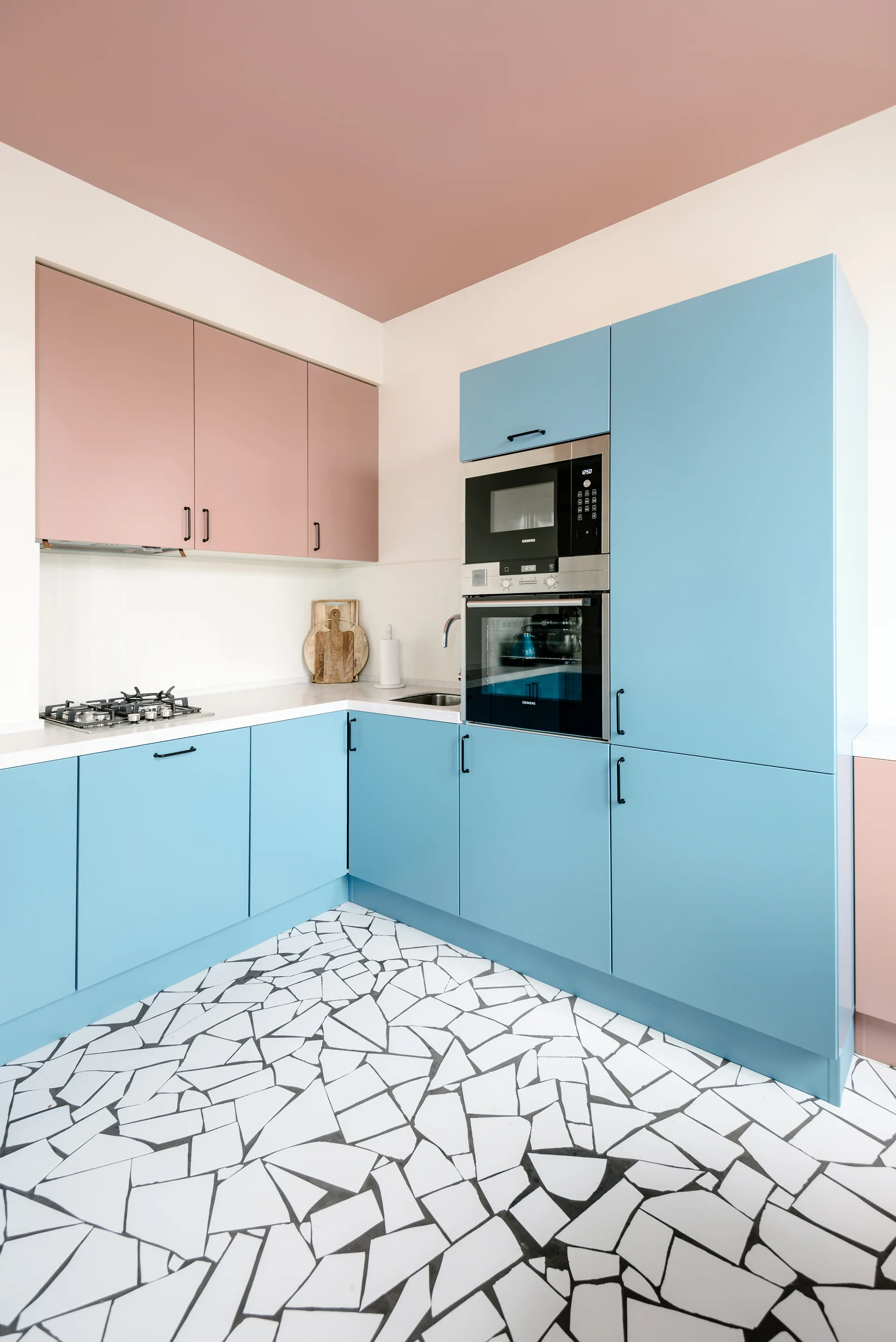 A modern kitchen featuring pastel blue and pink cabinets, a white backsplash, integrated appliances, and a bold black-and-white fractured tile floor.
