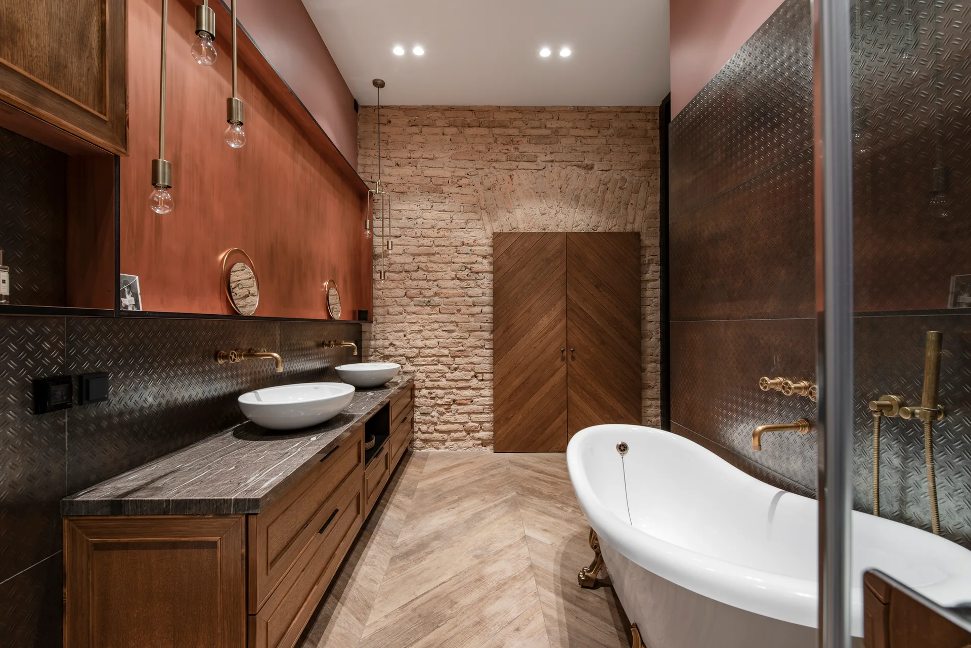 An industrial bathroom features a freestanding clawfoot tub, a double wooden vanity, exposed brick, and textured metal wall cladding.