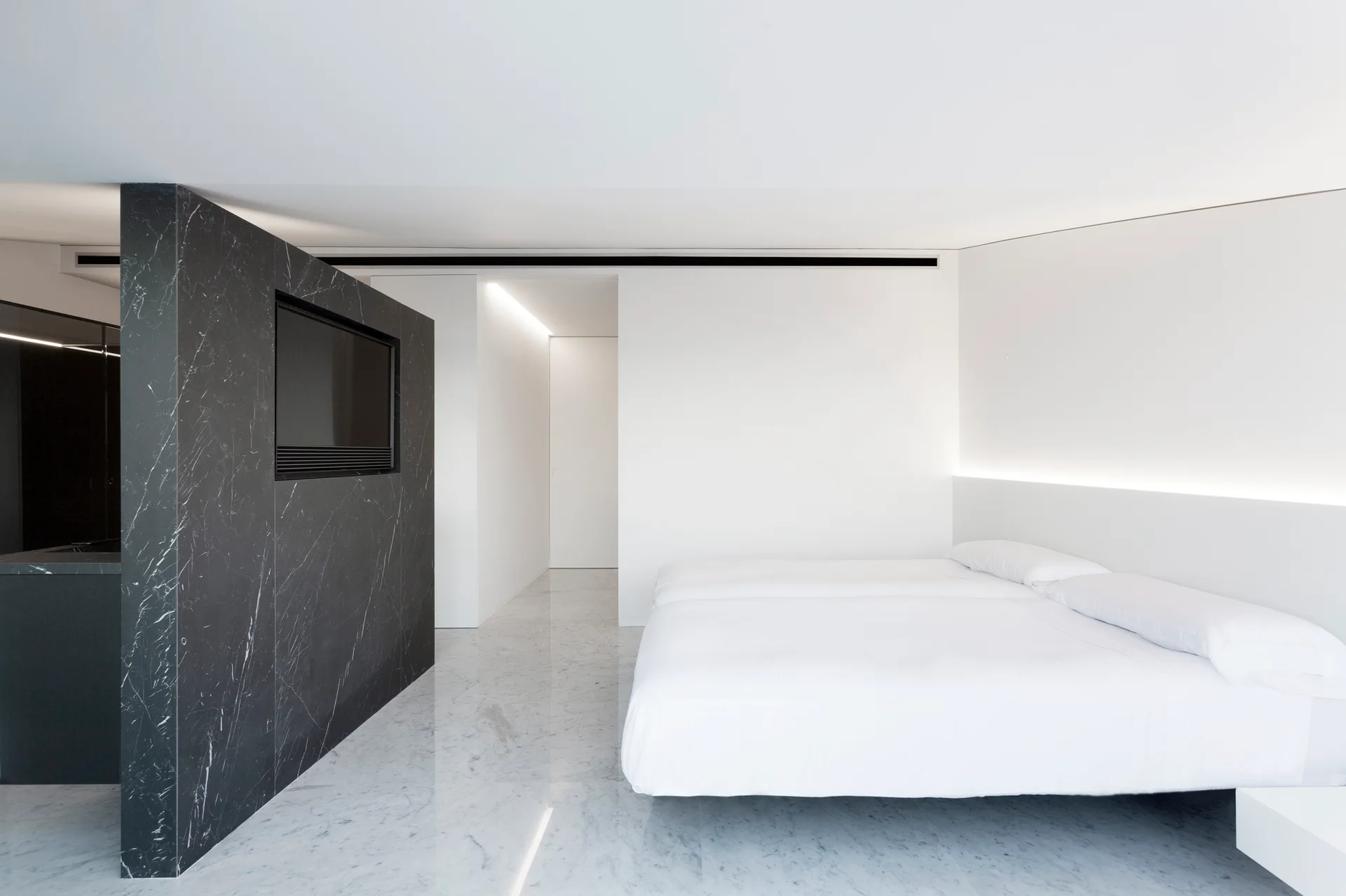A minimalist white bedroom featuring a large platform bed, polished white marble floors, and a dark marble partition wall with a built-in television screen.