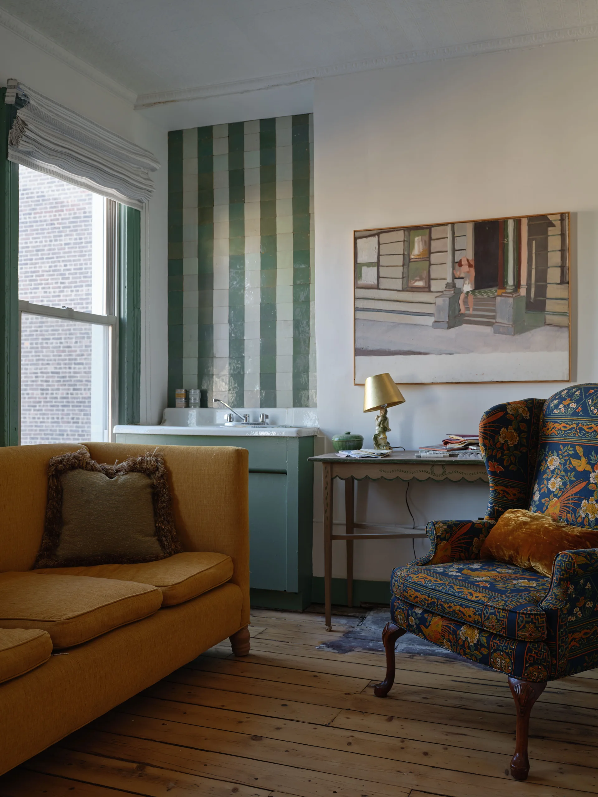 An eclectic living room corner featuring a yellow sofa, a floral wingback armchair, and a small sink backed by green and white striped tiles.
