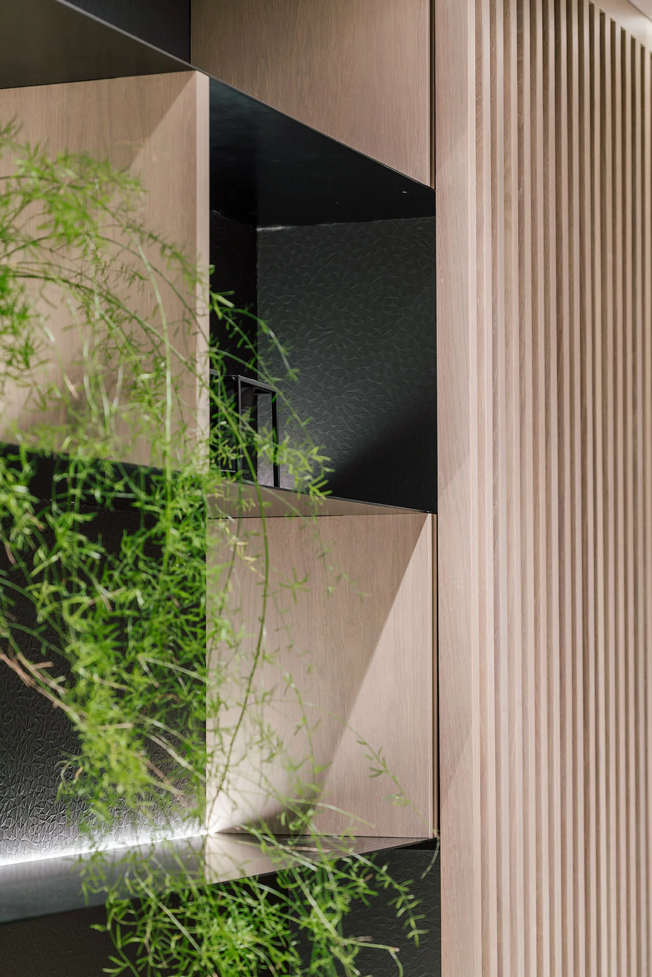 A close-up of built-in shelving featuring black recessed compartments, a textured dark back panel, warm integrated lighting, and vertical light-wood slats.