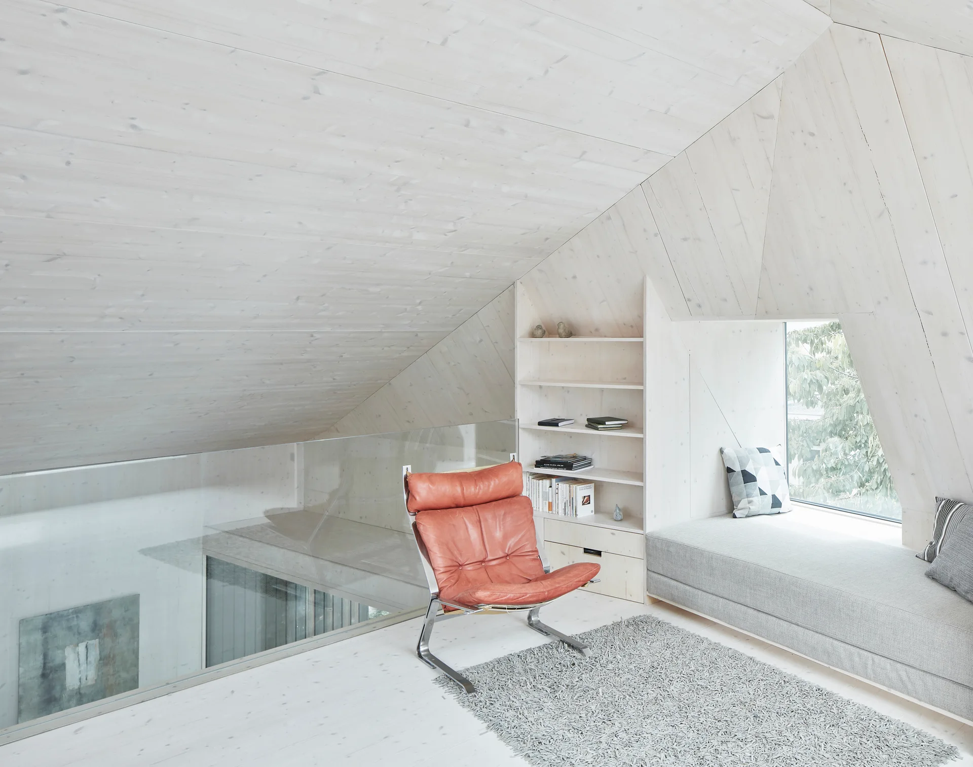 A bright attic landing features whitewashed wood paneling, a slanted ceiling, a cognac leather lounge chair, and a built-in window bench with gray cushions.
