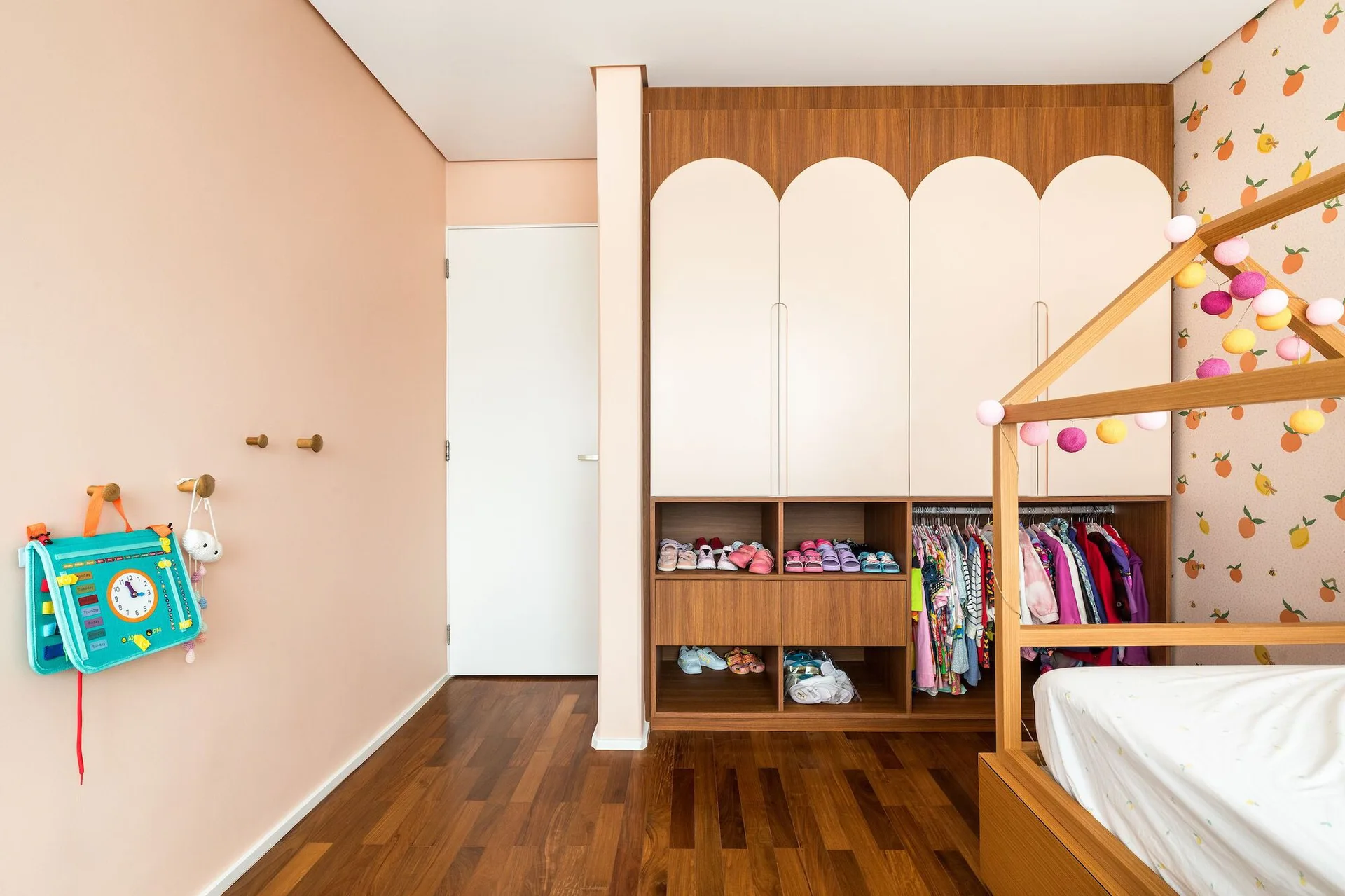 A children's bedroom features a wooden house-shaped bed frame, peach-colored walls, and a built-in wardrobe with arched peach-and-wood doors.