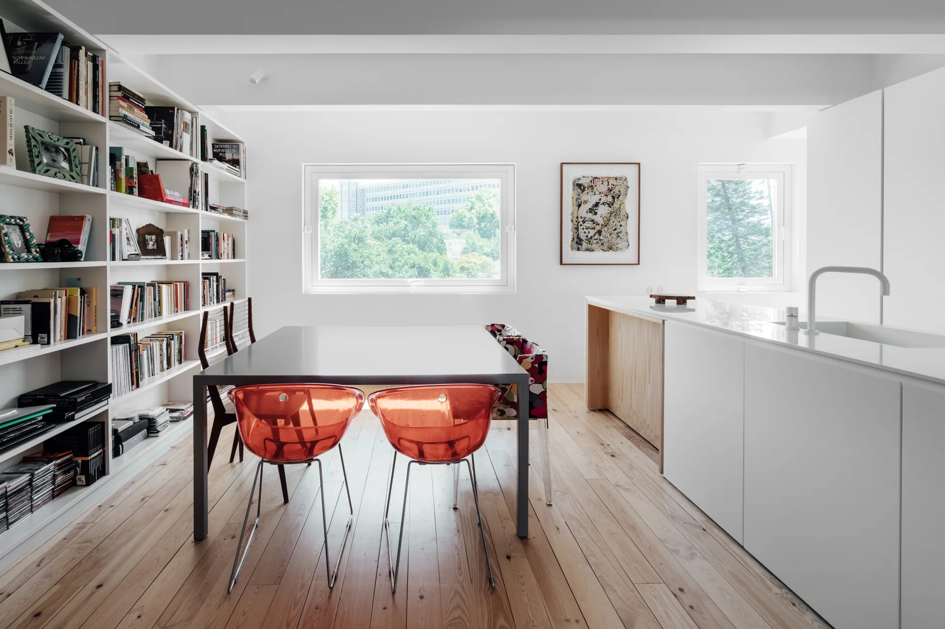 A minimalist dining area features a dark wood table with translucent orange chairs next to a large white bookcase and a kitchen island.