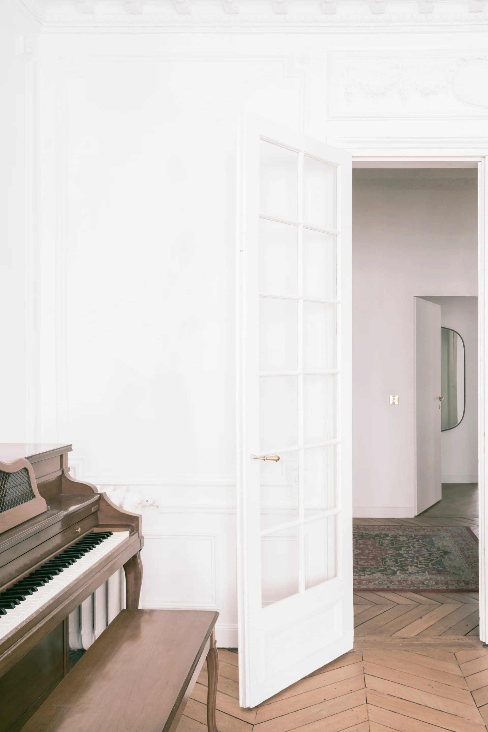 A wooden upright piano sits on a herringbone hardwood floor next to a white glass-paneled French door in a bright room.