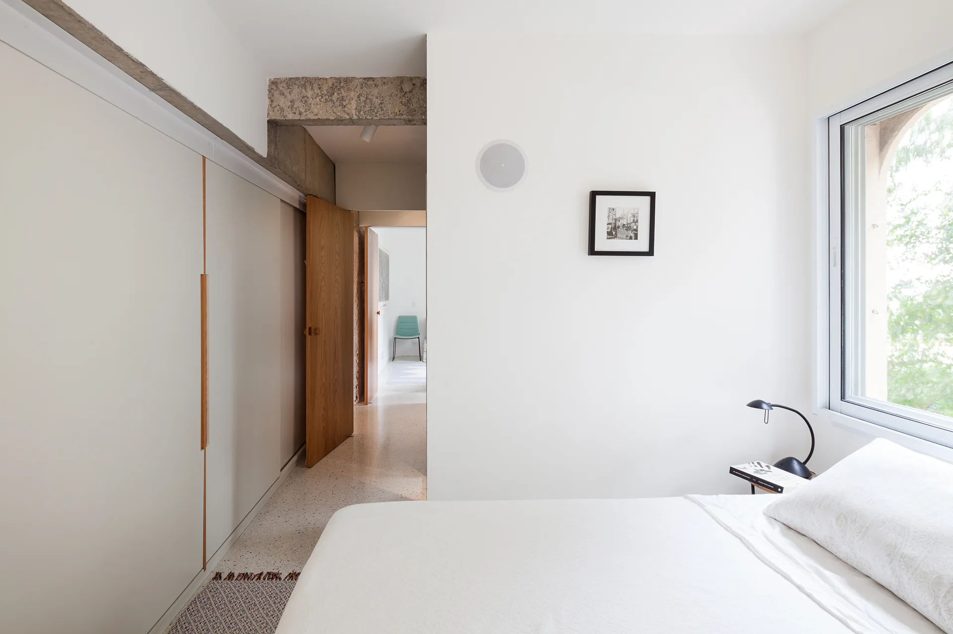 A minimalist white bedroom features a bed with light bedding, a window, built-in wardrobes, speckled terrazzo flooring, and an exposed concrete beam.