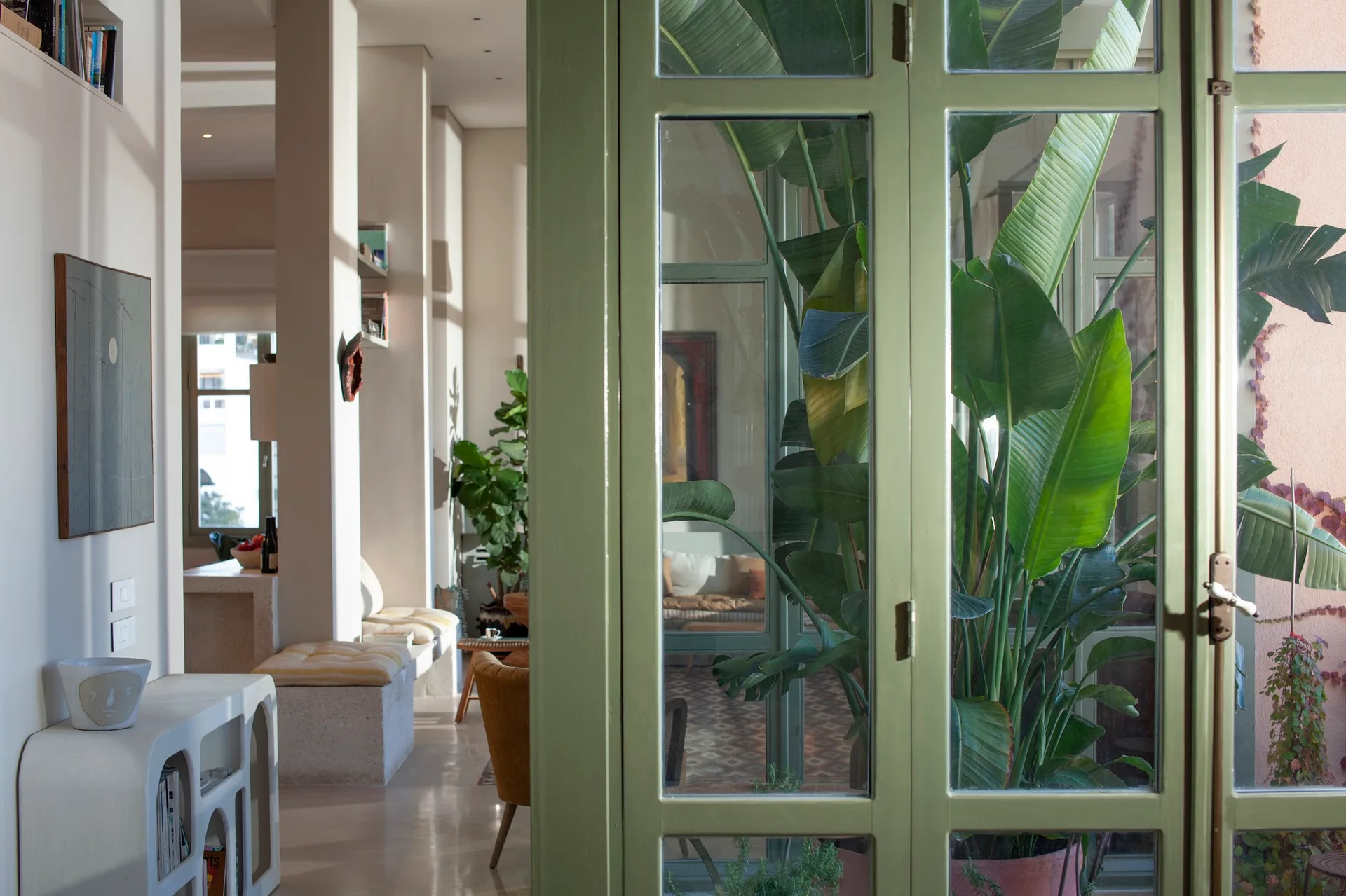A bright living space with a built-in bench and arched console is seen through a sage green glass door next to a large tropical plant.