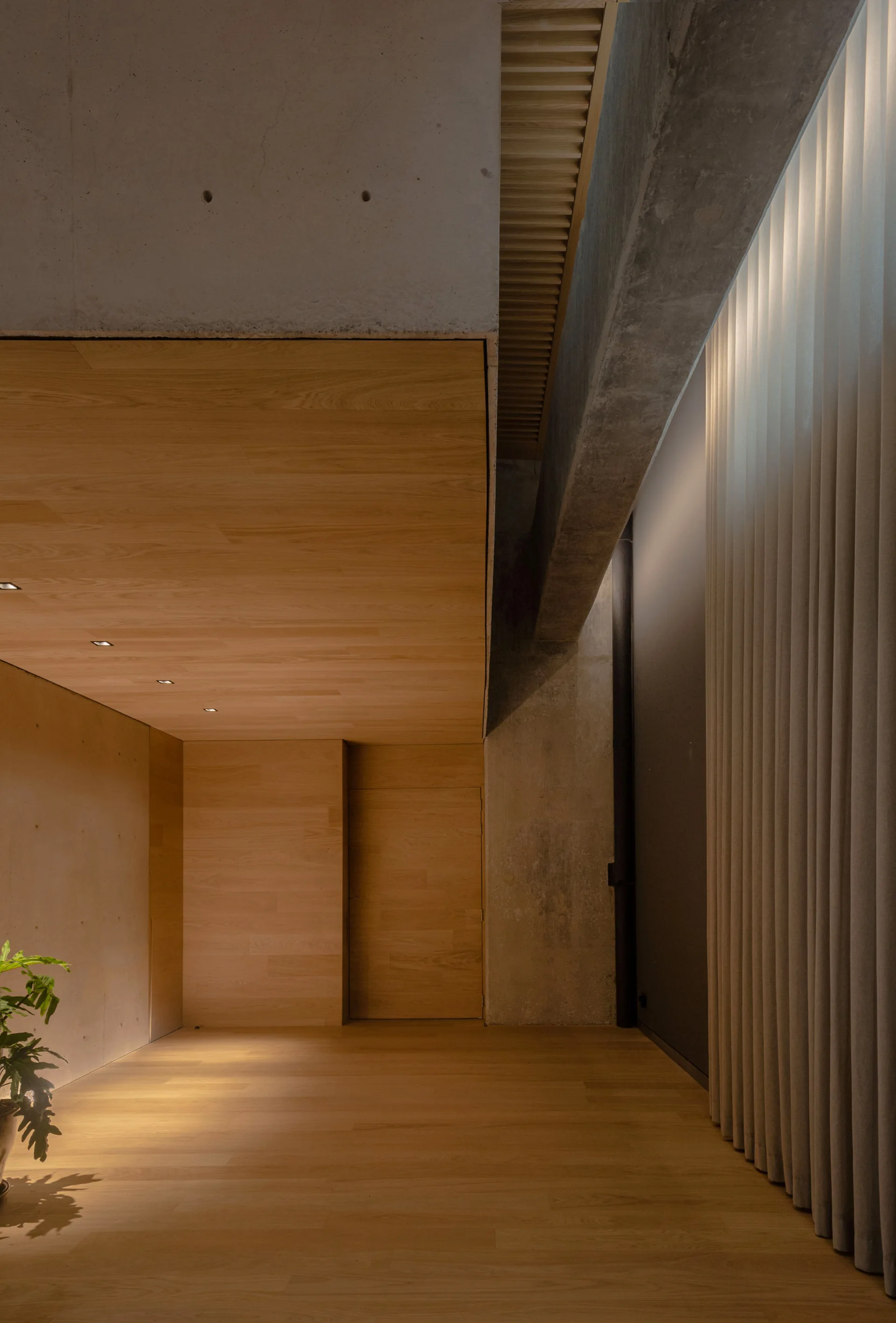 A minimalist hallway featuring warm oak wood paneling, an integrated flush door, a raw concrete structural beam, and tall pleated curtains.