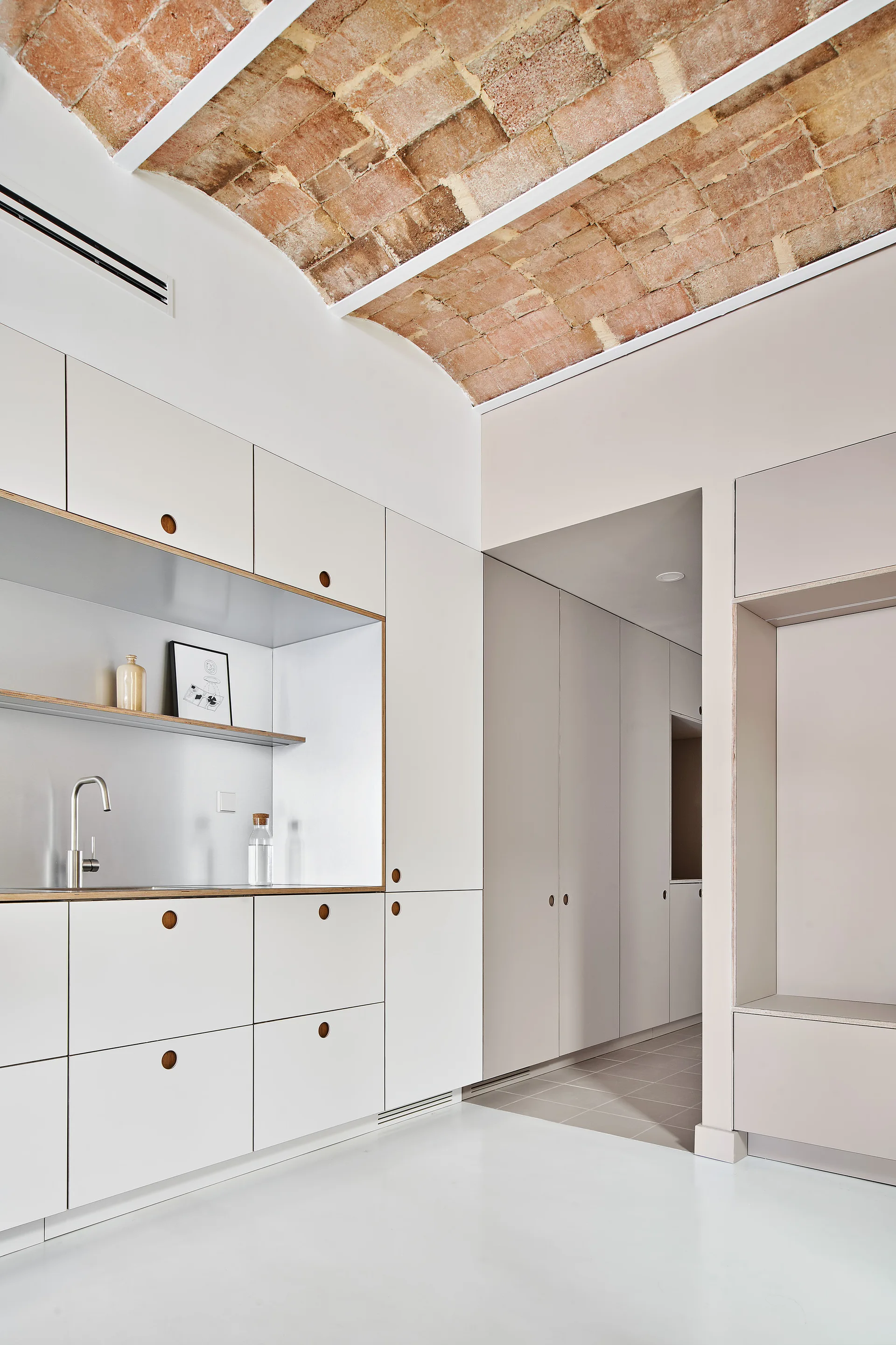 Minimalist off-white kitchen cabinetry features a recessed sink niche beneath a rustic vaulted brick ceiling.