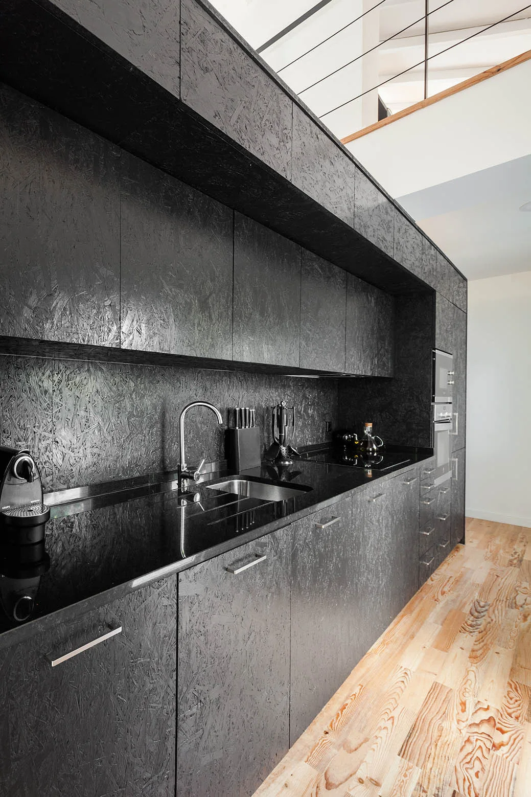 A modern kitchen features textured black OSB cabinets, a polished black countertop, and light wood plank flooring.