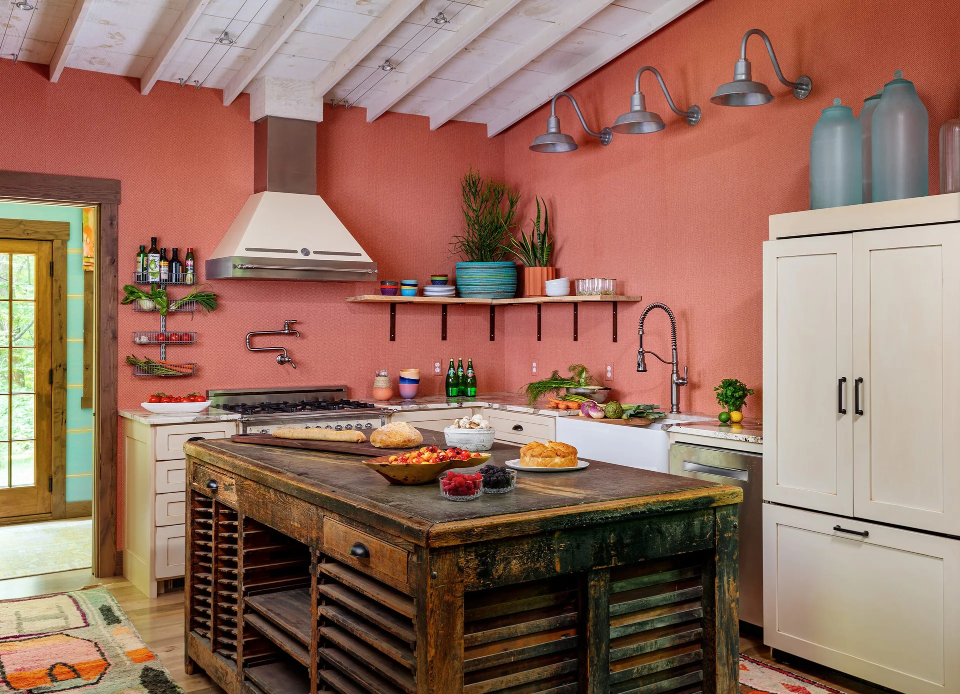 A rustic kitchen features a distressed dark wood island, textured salmon-pink walls, cream-colored appliances, and a white wood-beamed ceiling.