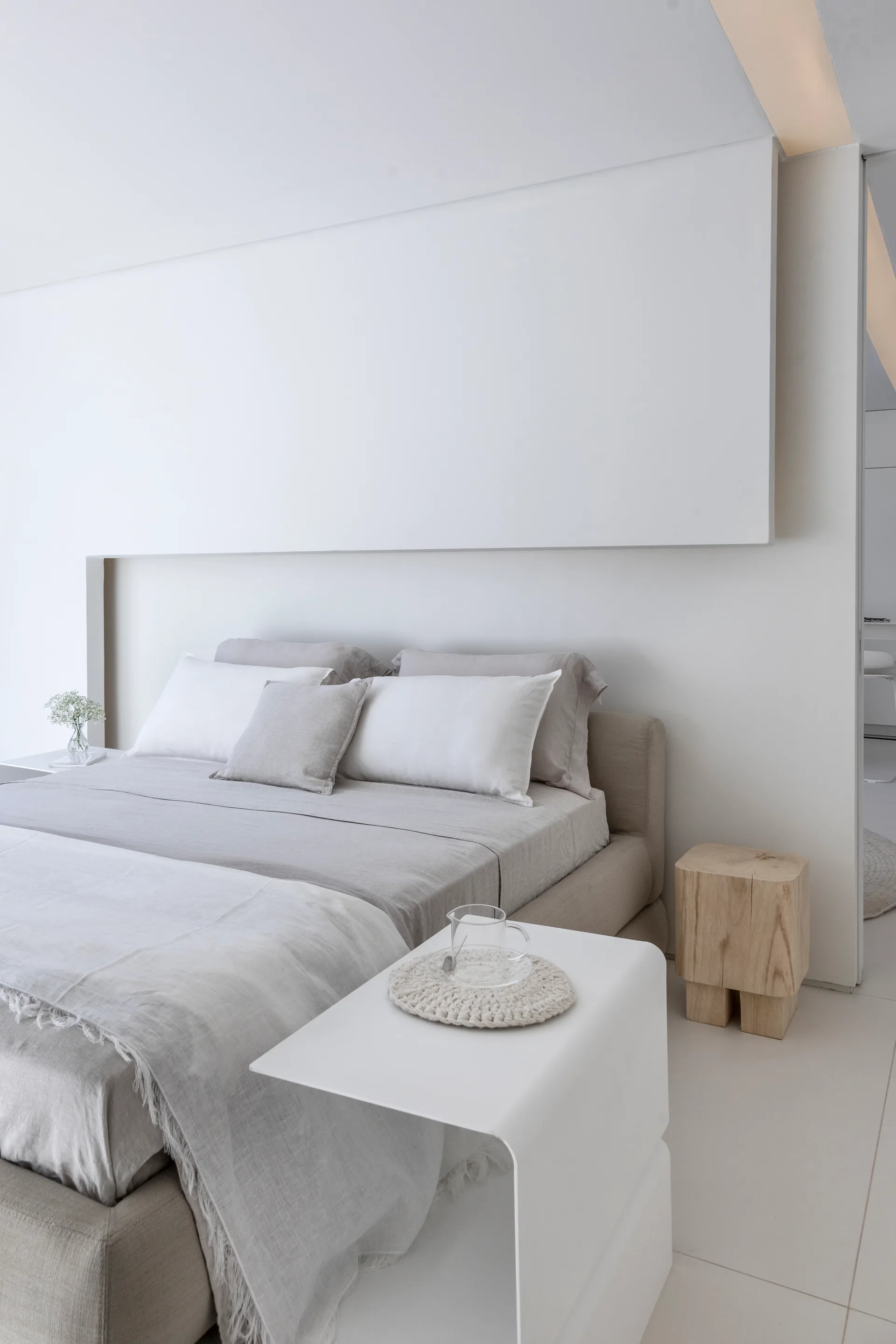 A minimalist bedroom features a beige upholstered bed with linen bedding, a curved white bedside table, and a solid wood block stool.