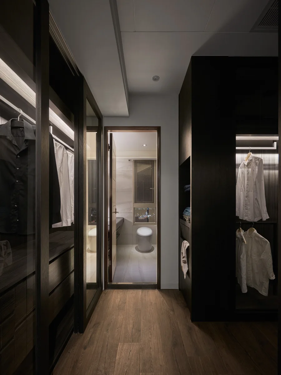 A modern walk-in closet with dark wood wardrobes and integrated lighting leads to an adjacent bathroom with a visible toilet.