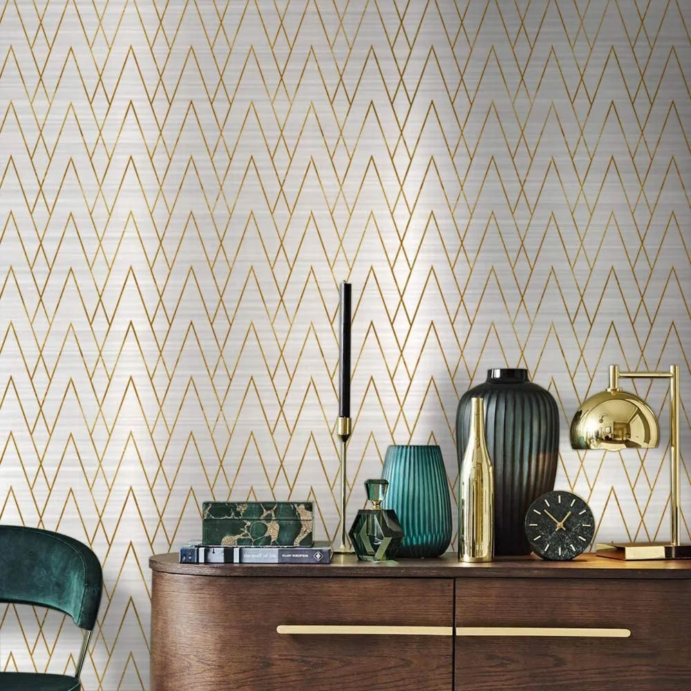 A dark wood sideboard topped with a brass dome lamp and green glass vases sits against a white and gold geometric patterned wallpaper.