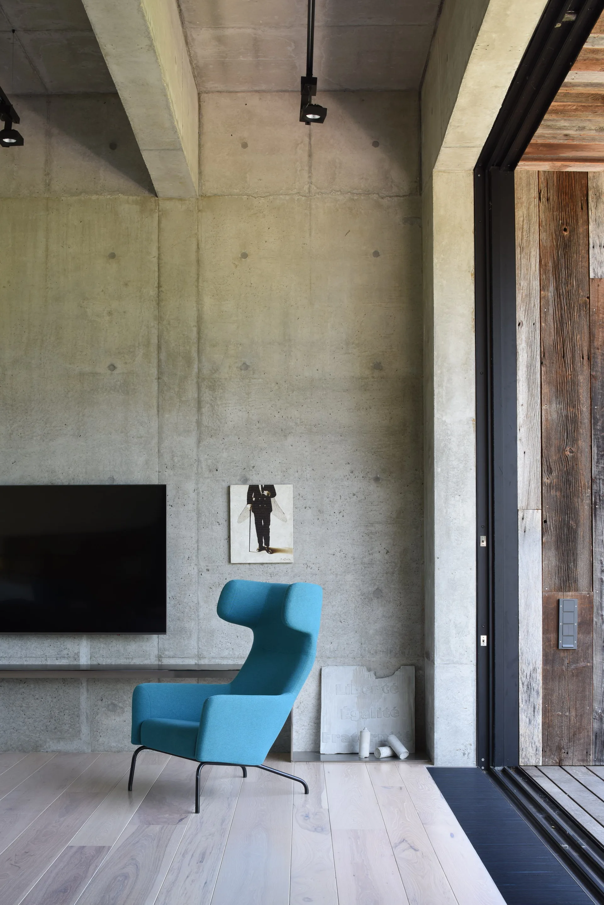 A bright blue armchair sits on light wood flooring against an exposed concrete wall next to a large sliding glass door.