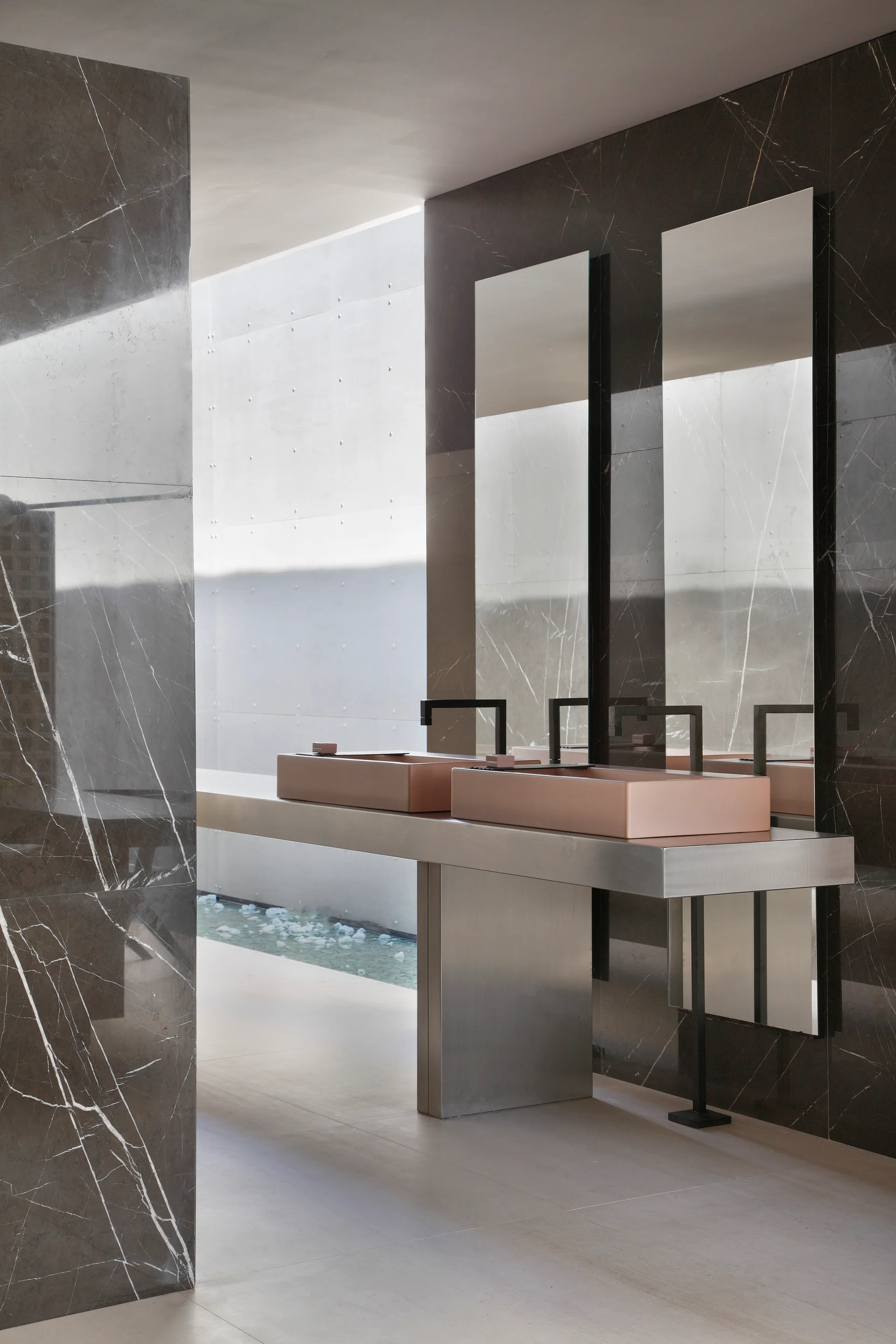 A modern bathroom features a brushed steel vanity console with two rectangular rose gold sinks against dark veined marble walls.
