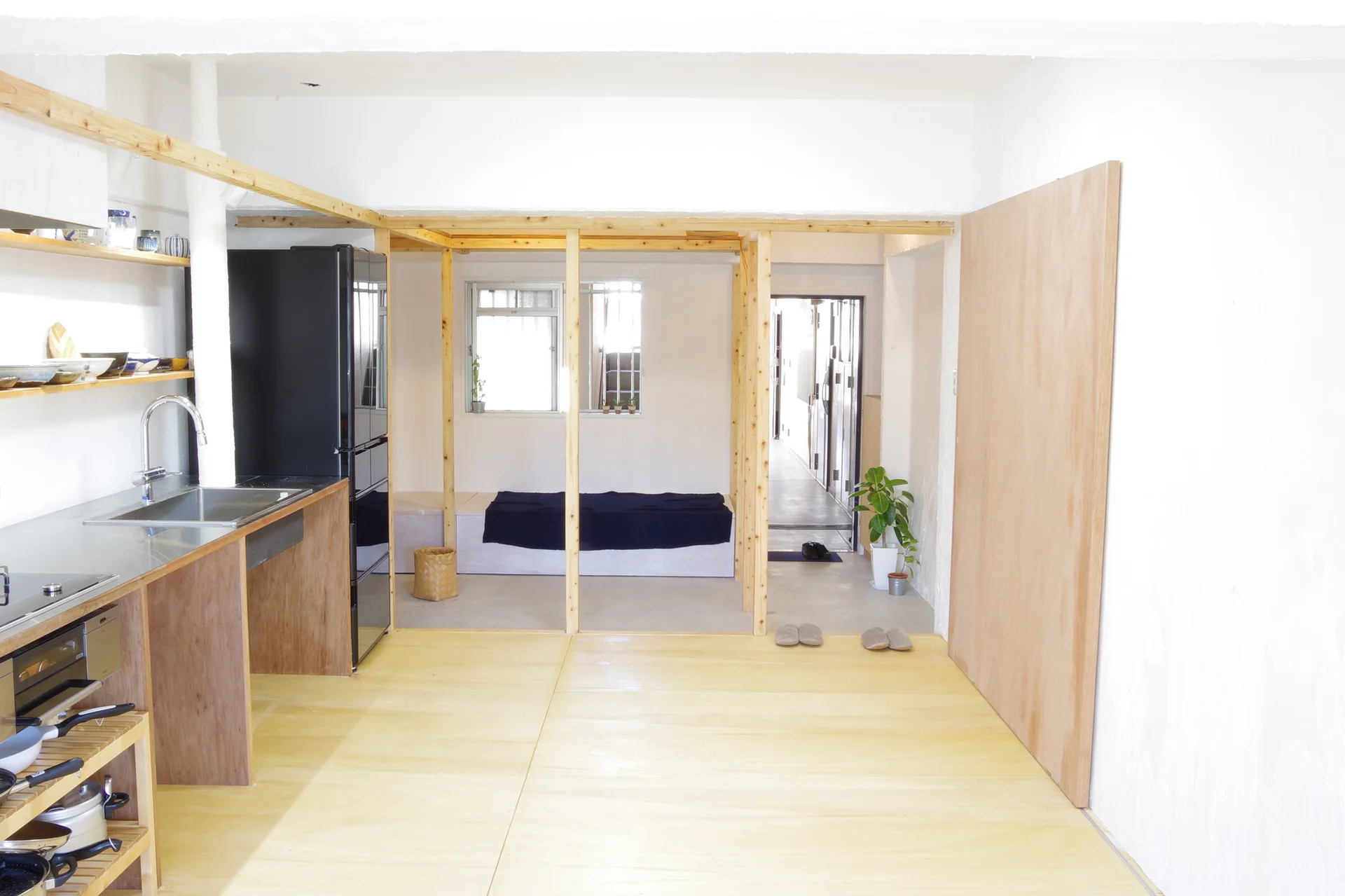 A minimalist kitchen features plywood flooring, a stainless steel sink counter, and an exposed light wood timber frame.