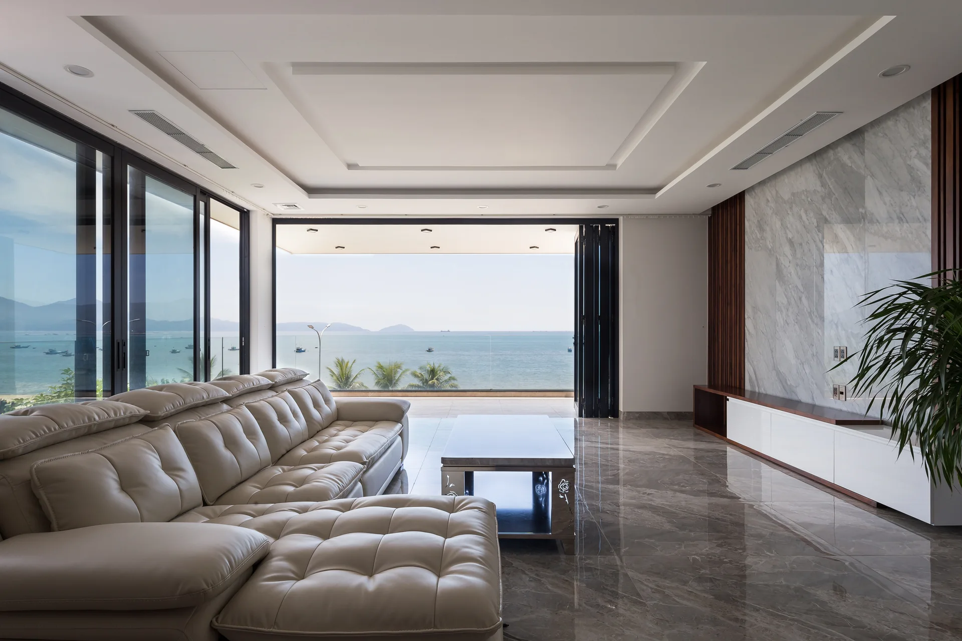 A contemporary living room features a cream leather sectional sofa, polished grey marble floors, and sliding glass doors that open to an ocean view.