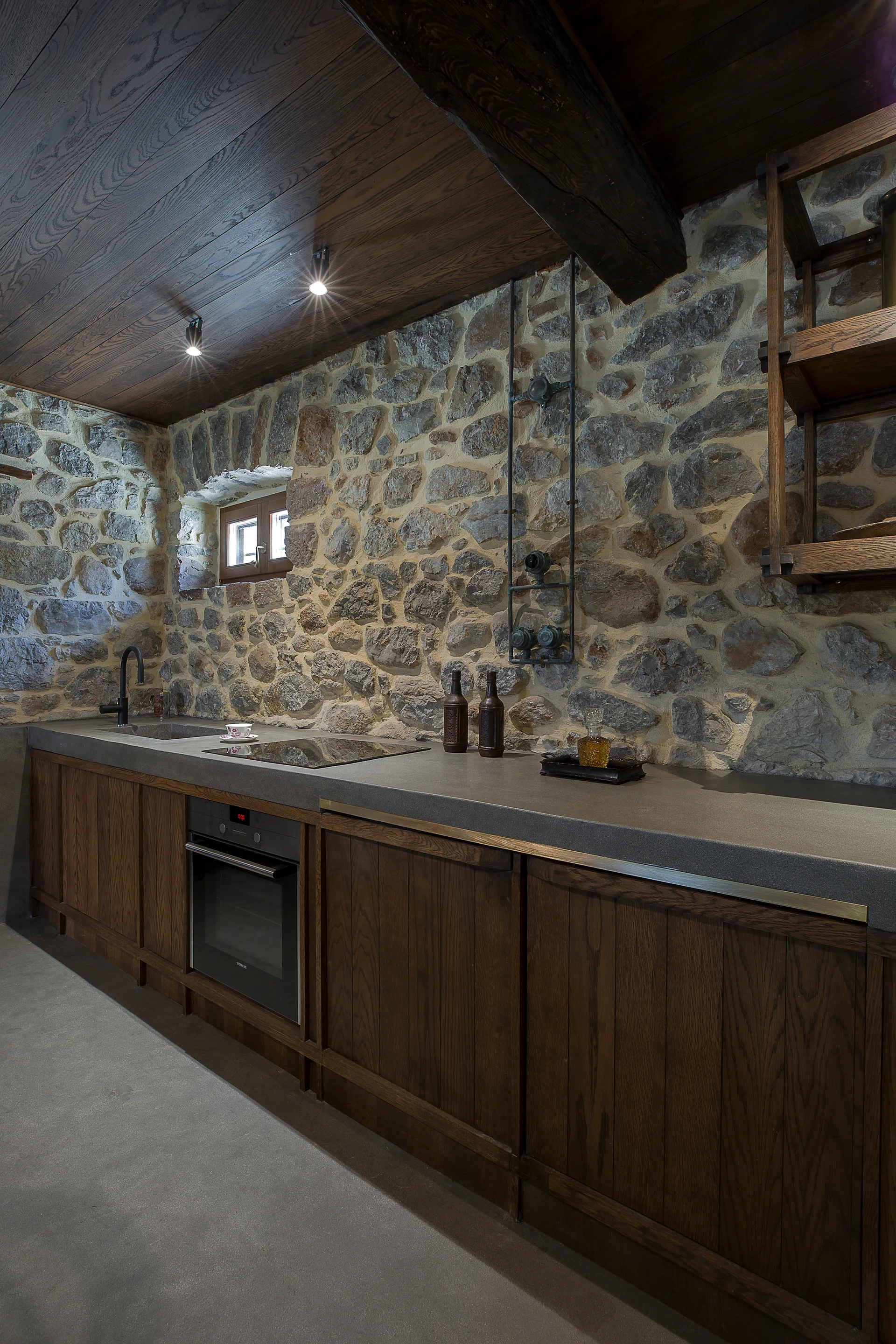 A rustic kitchen featuring dark wood cabinets, a thick grey countertop, and a textured stone wall under a dark wooden plank ceiling.