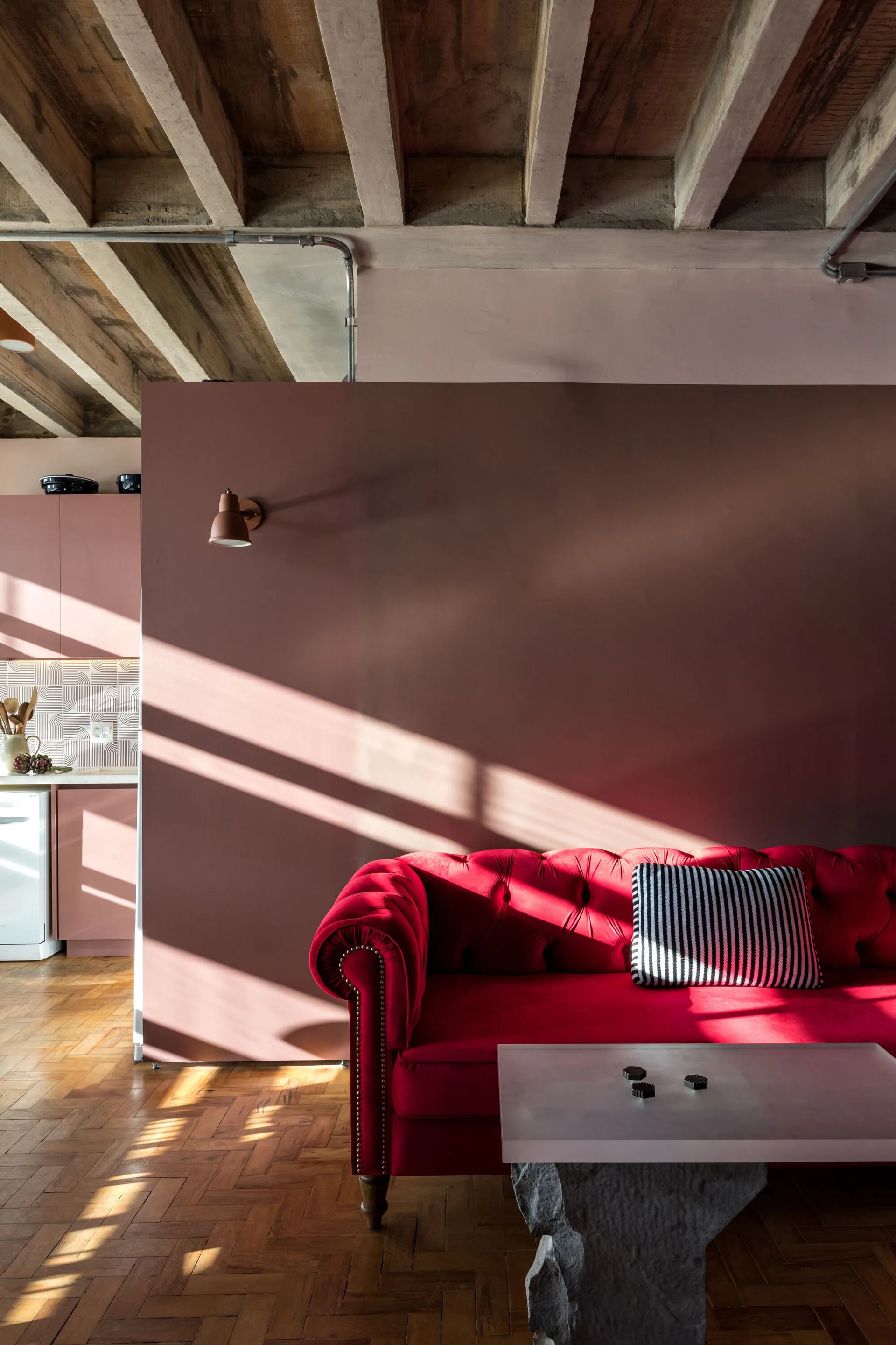 A tufted red velvet sofa sits against a mauve partition wall under exposed ceiling beams in a living room with herringbone parquet wood floors.