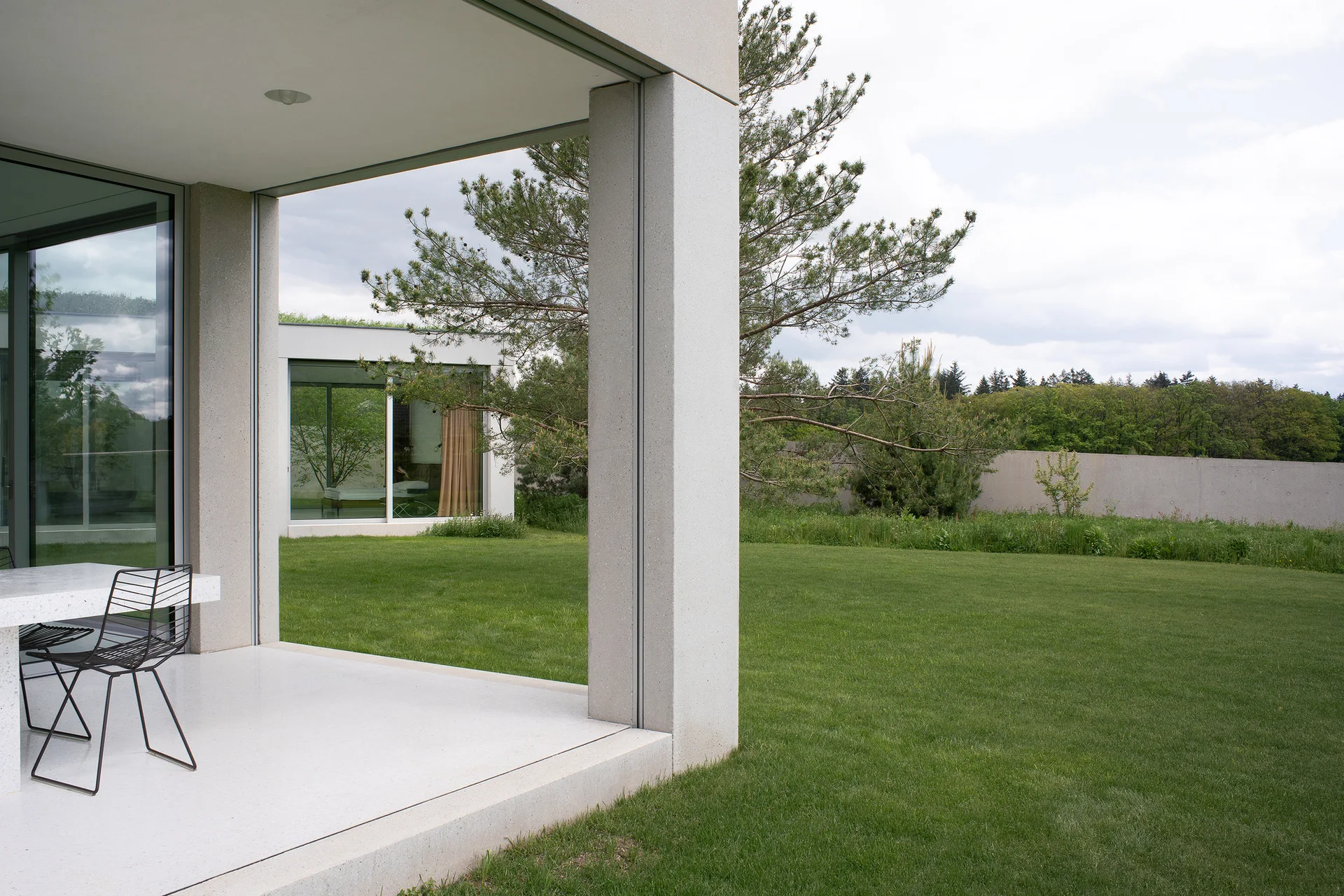 A minimalist covered concrete patio with a white terrazzo table and black wire chairs looks out onto a manicured green lawn.