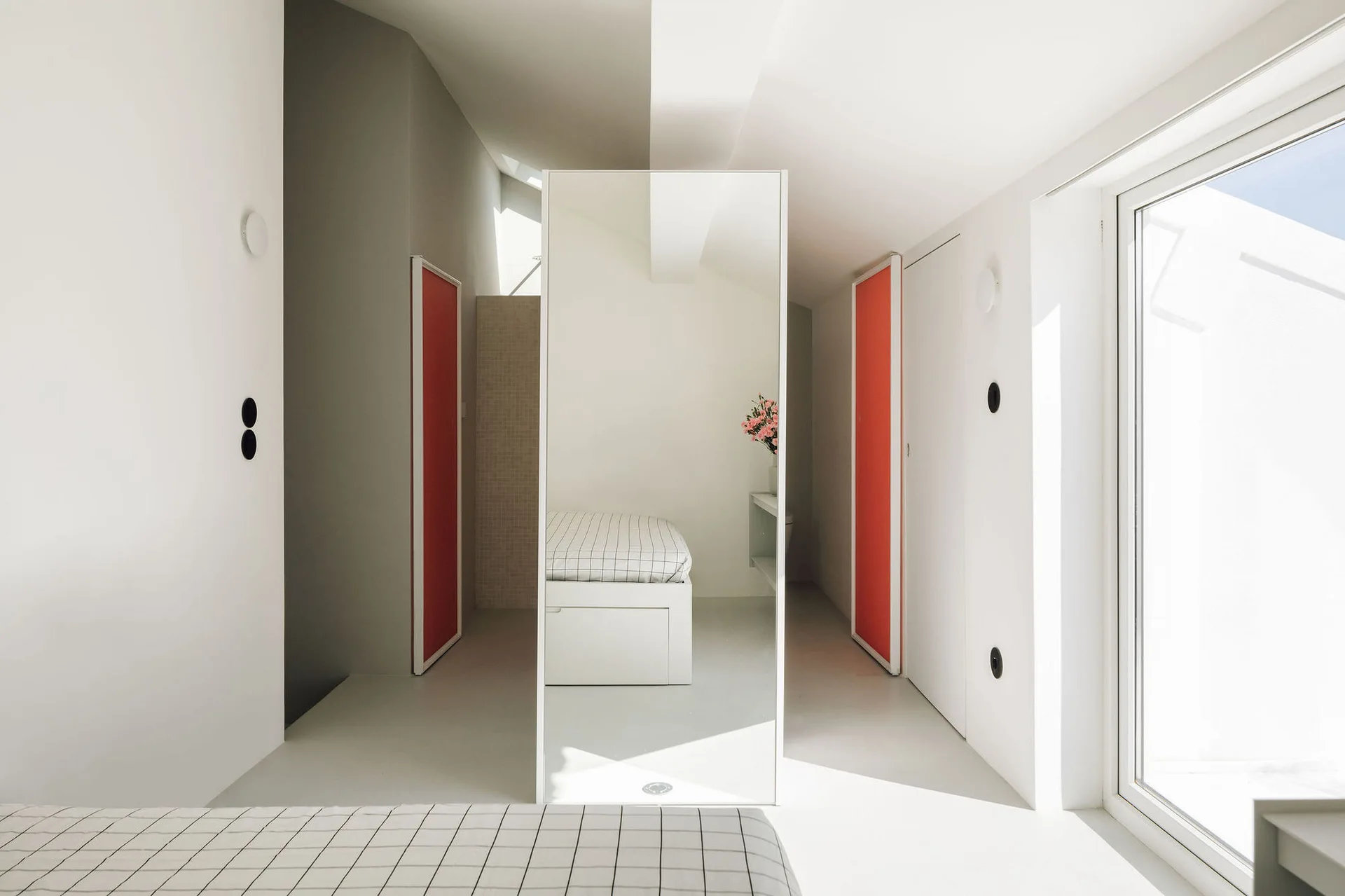 A minimalist white bedroom featuring a central full-length mirror and two sliding doors painted in bright orange.