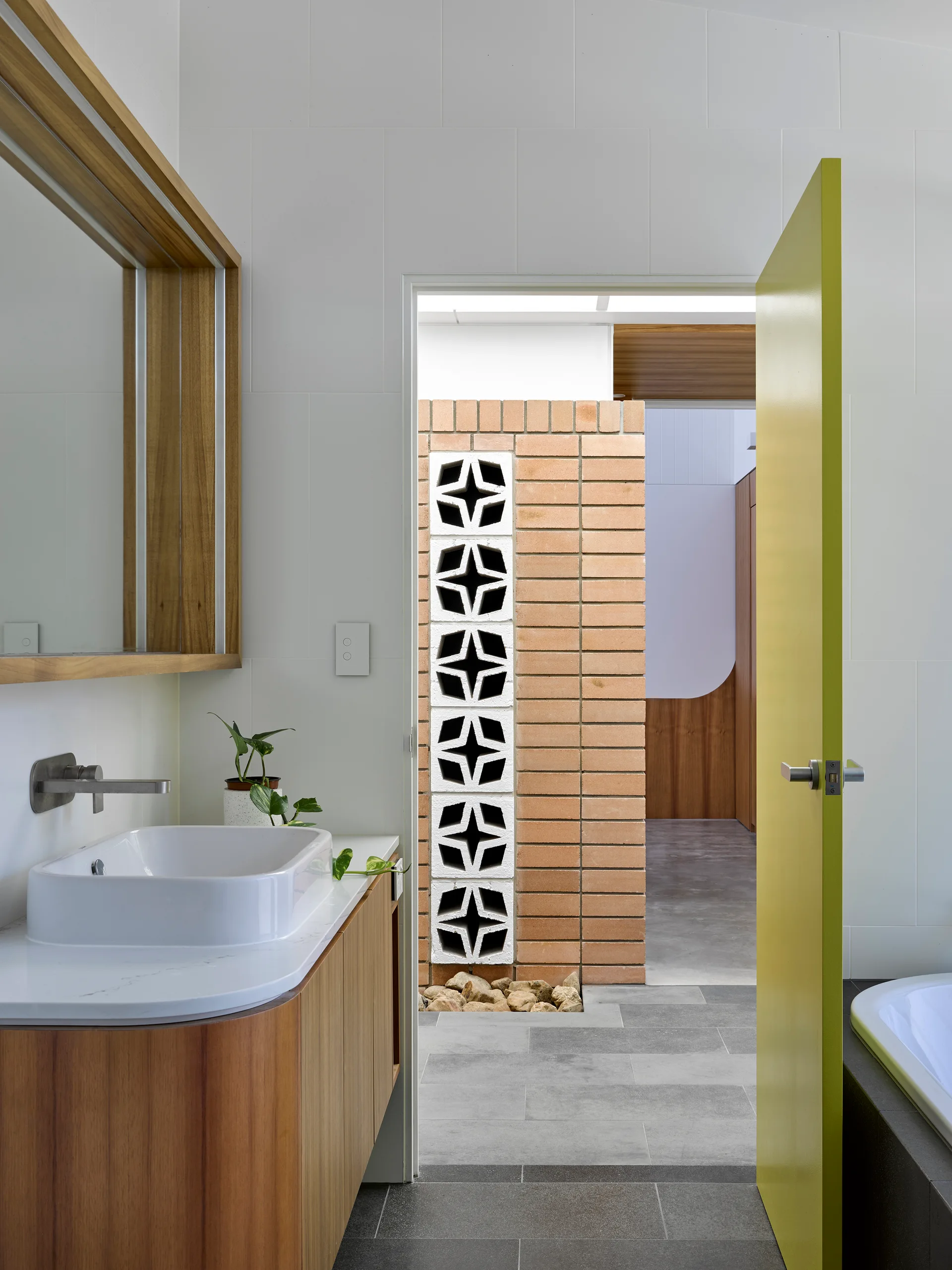 A modern bathroom features a wood vanity and a bright chartreuse door open to a brick courtyard with decorative breeze blocks.