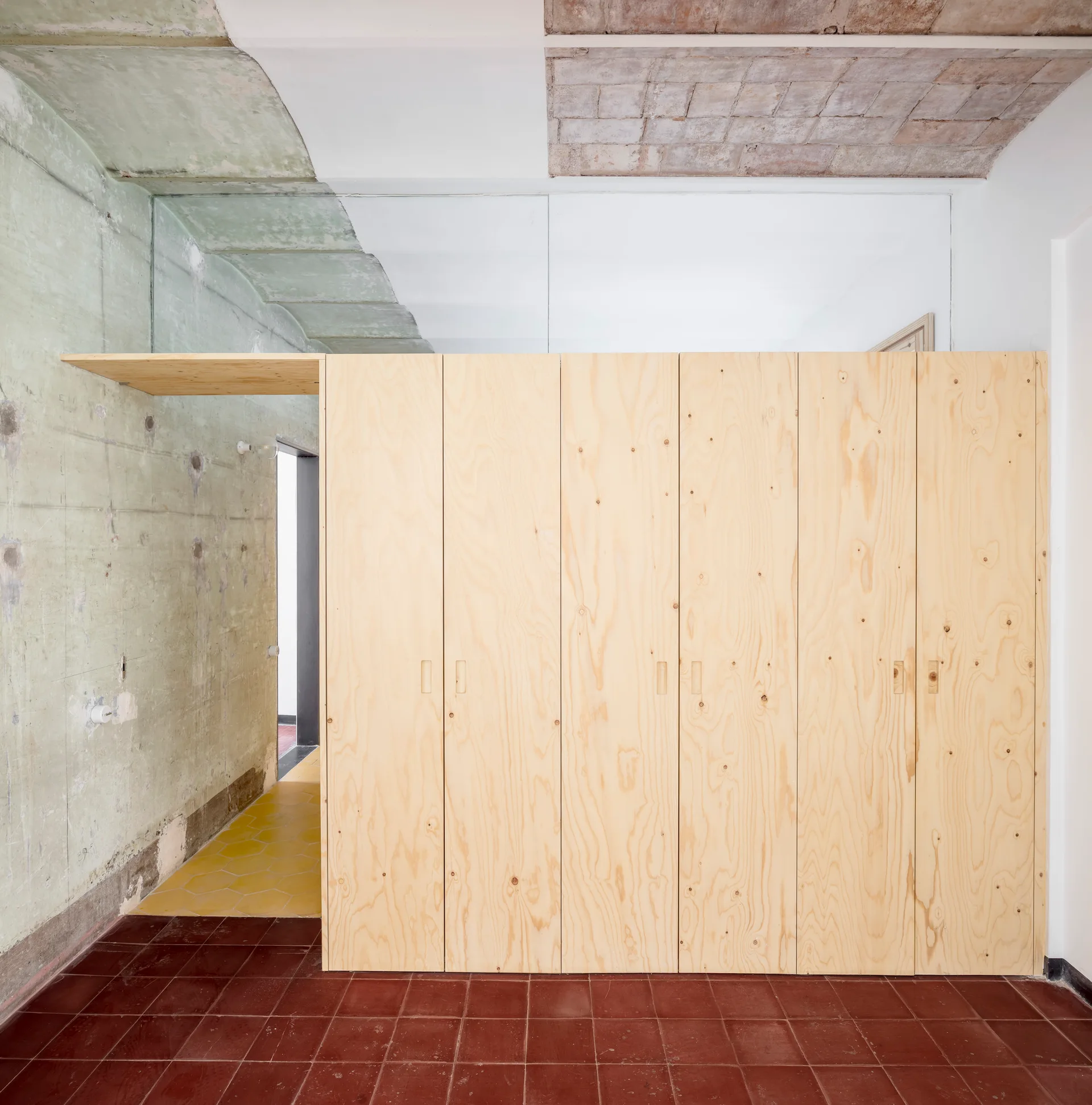 A large minimalist plywood built-in closet stands on red terracotta tile flooring next to a raw concrete wall.