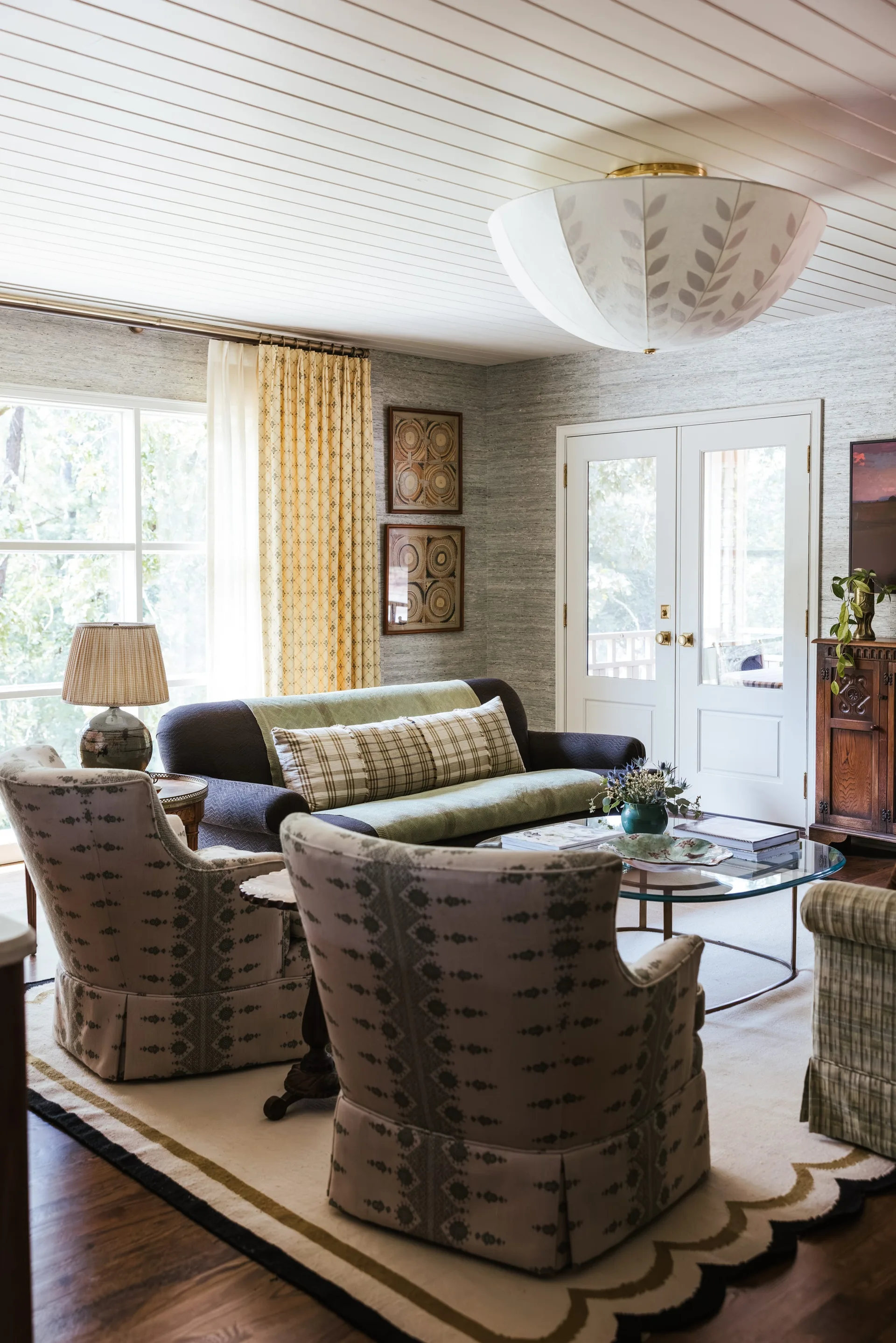 A cozy living room features patterned armchairs, a glass coffee table, grey grasscloth walls, and a white planked ceiling.