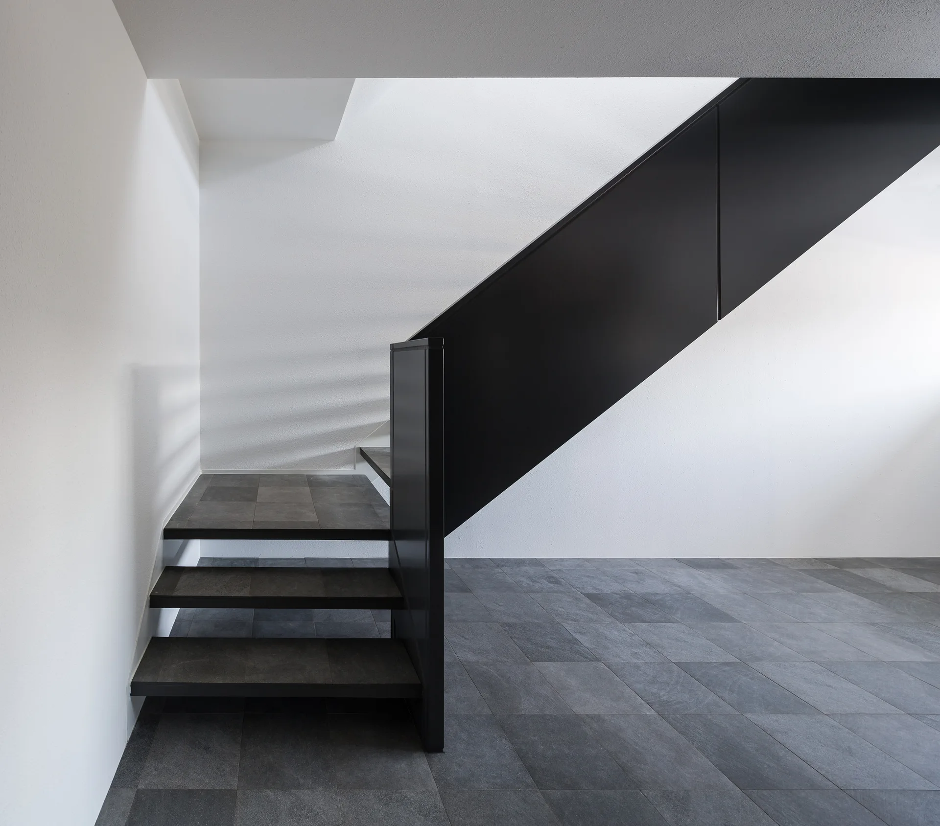 A minimalist black metal staircase with dark stone-like tiled steps stands against plain white walls and a matching dark tiled floor.