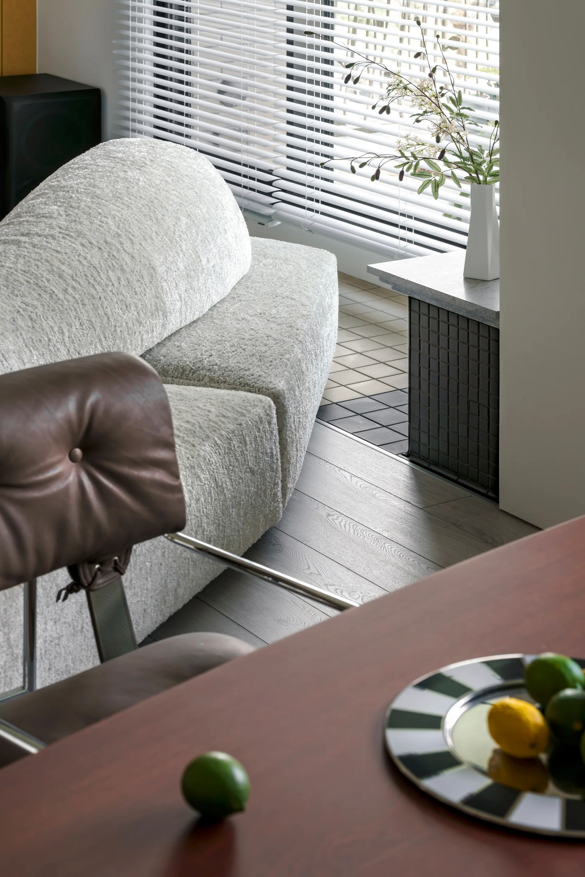 A white textured boucle sofa sits near window blinds, next to a tiled side table and a wood dining table with a striped platter.