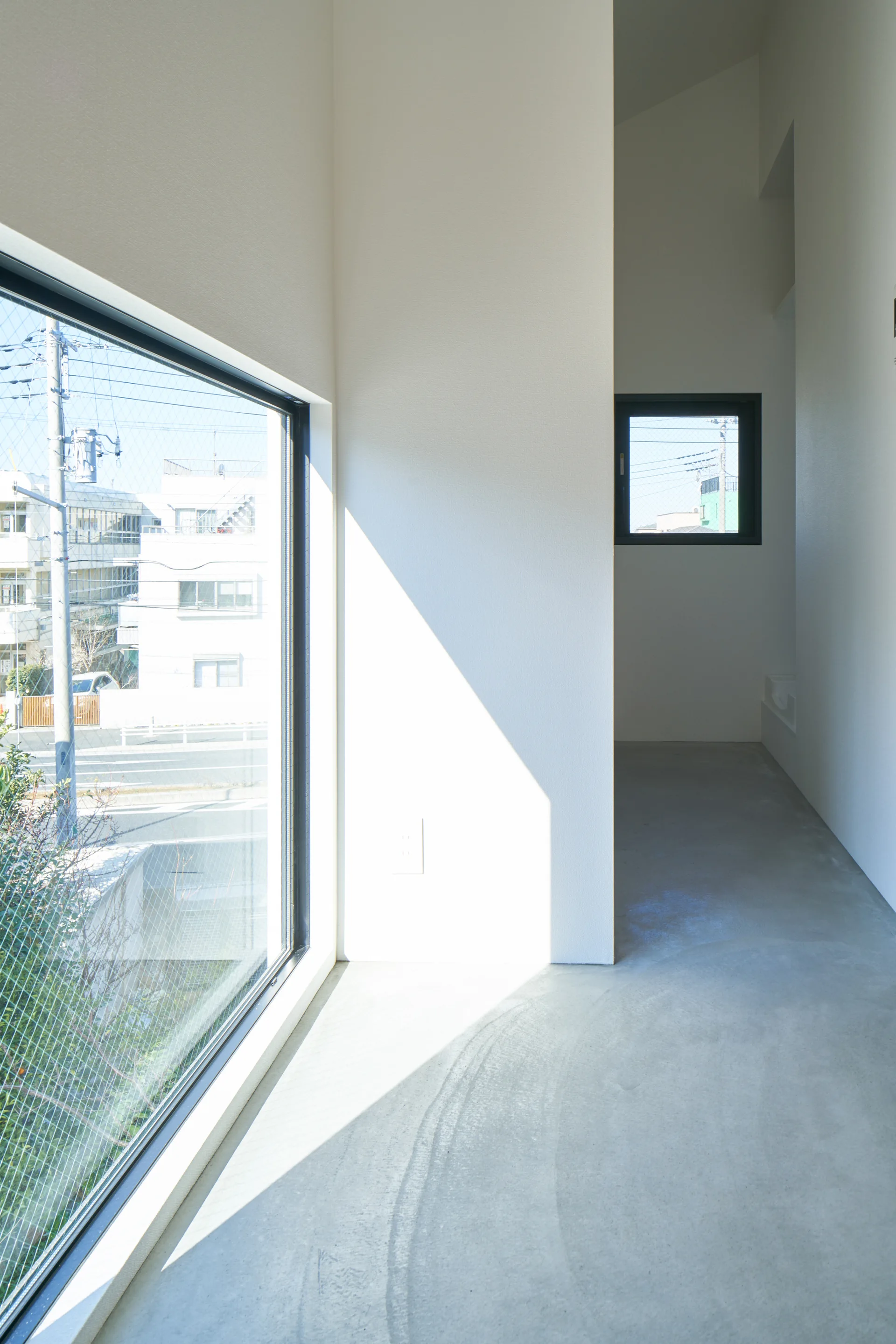 A minimalist hallway features smooth light grey concrete floors, plain white walls, and a large black-framed window letting in bright natural light.