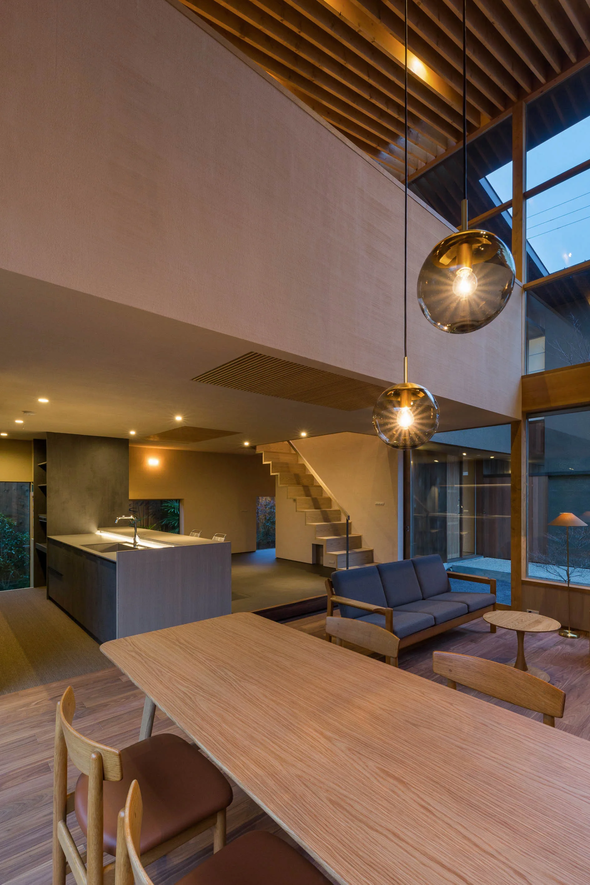 An open plan modern interior featuring a wood dining table, gray sofa, spherical pendant lights, and an open staircase beneath exposed ceiling rafters.