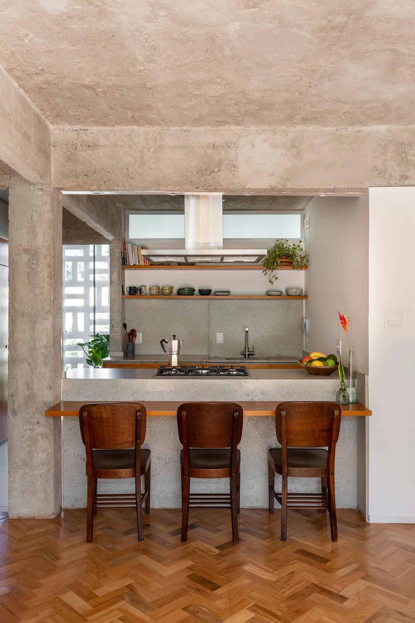 Three wooden bar stools stand at a gray stone island under an exposed concrete ceiling with herringbone wood flooring.