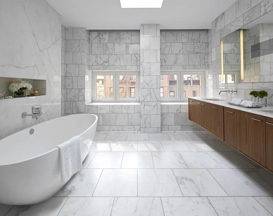 A spacious bathroom features floor-to-ceiling white marble tiles, a white freestanding soaking tub, and a long floating wood vanity.