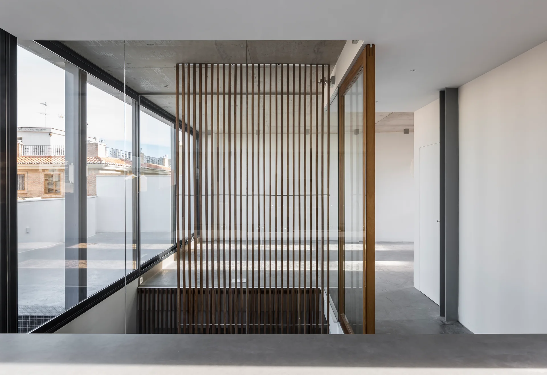 A minimalist modern interior featuring a vertical slatted wood room divider, concrete ceiling, and large glass windows.