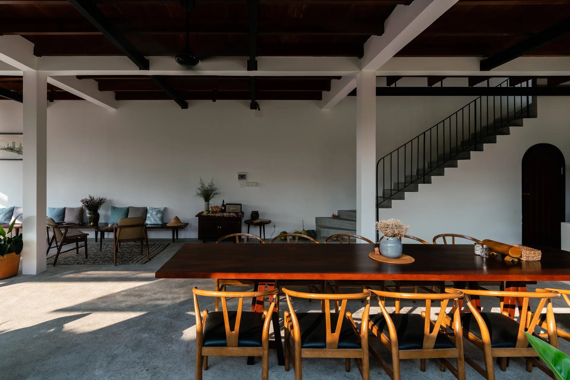 A long dark wood dining table surrounded by wooden chairs sits on a concrete floor beneath exposed dark ceiling beams in an open living space.