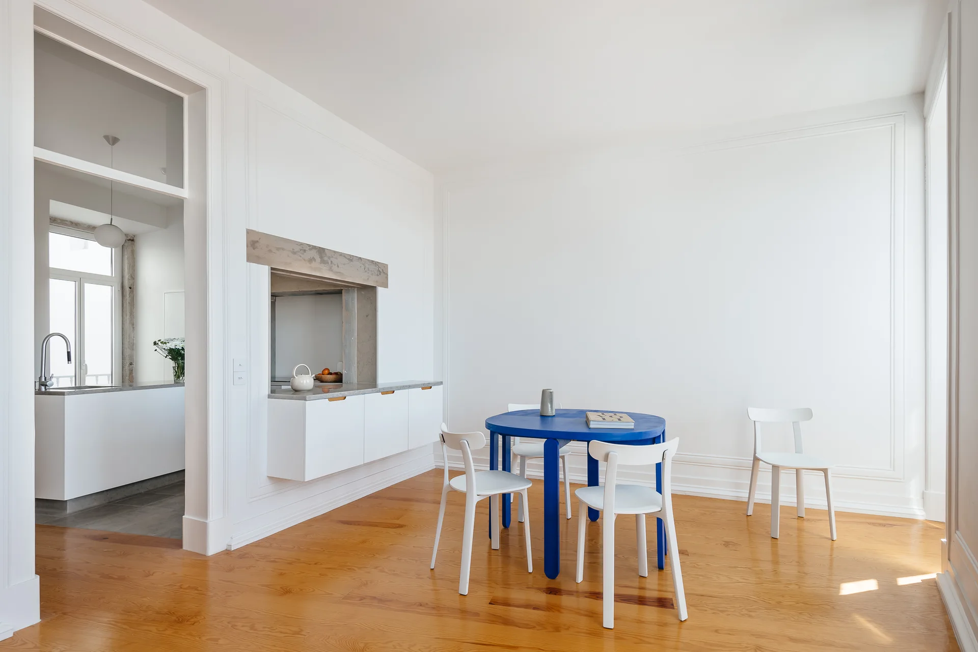 A bright dining area features a bold blue circular table, white dining chairs, light hardwood floors, and clean white walls.