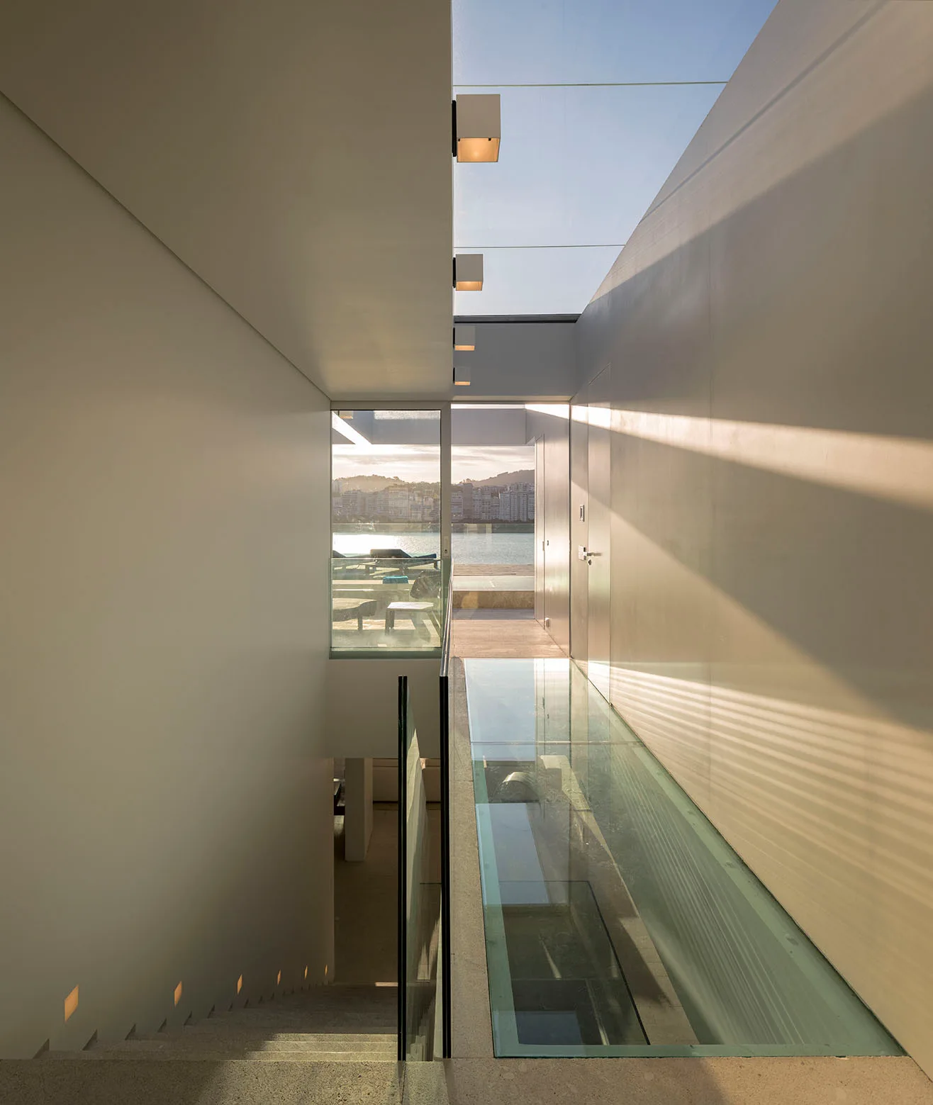 A modern minimalist staircase landing features a transparent glass walkway alongside stone steps, with a large window displaying a coastal view.
