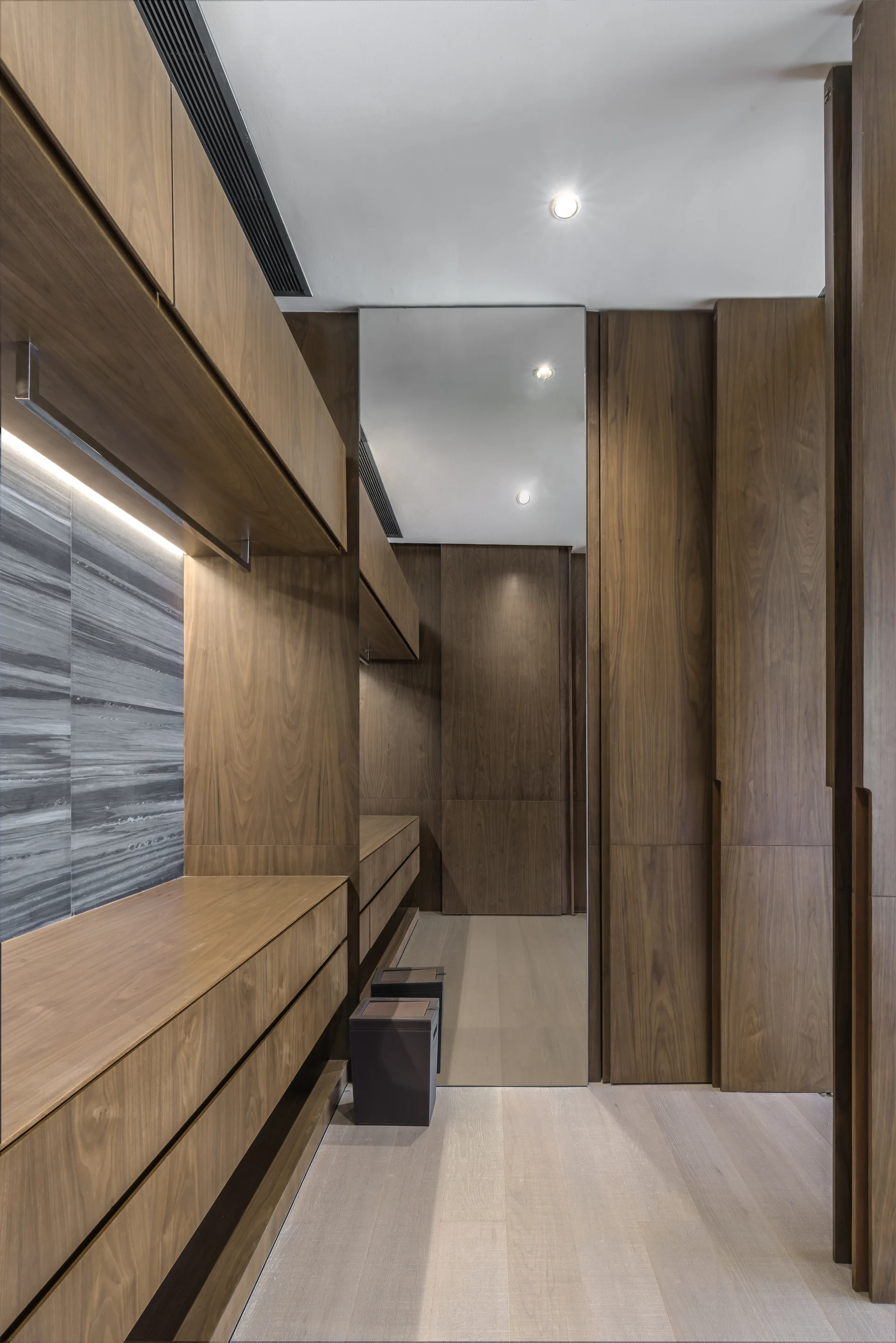A modern walk-in closet featuring warm wood cabinetry, a full-length wall mirror, light wood floors, and integrated under-cabinet lighting.