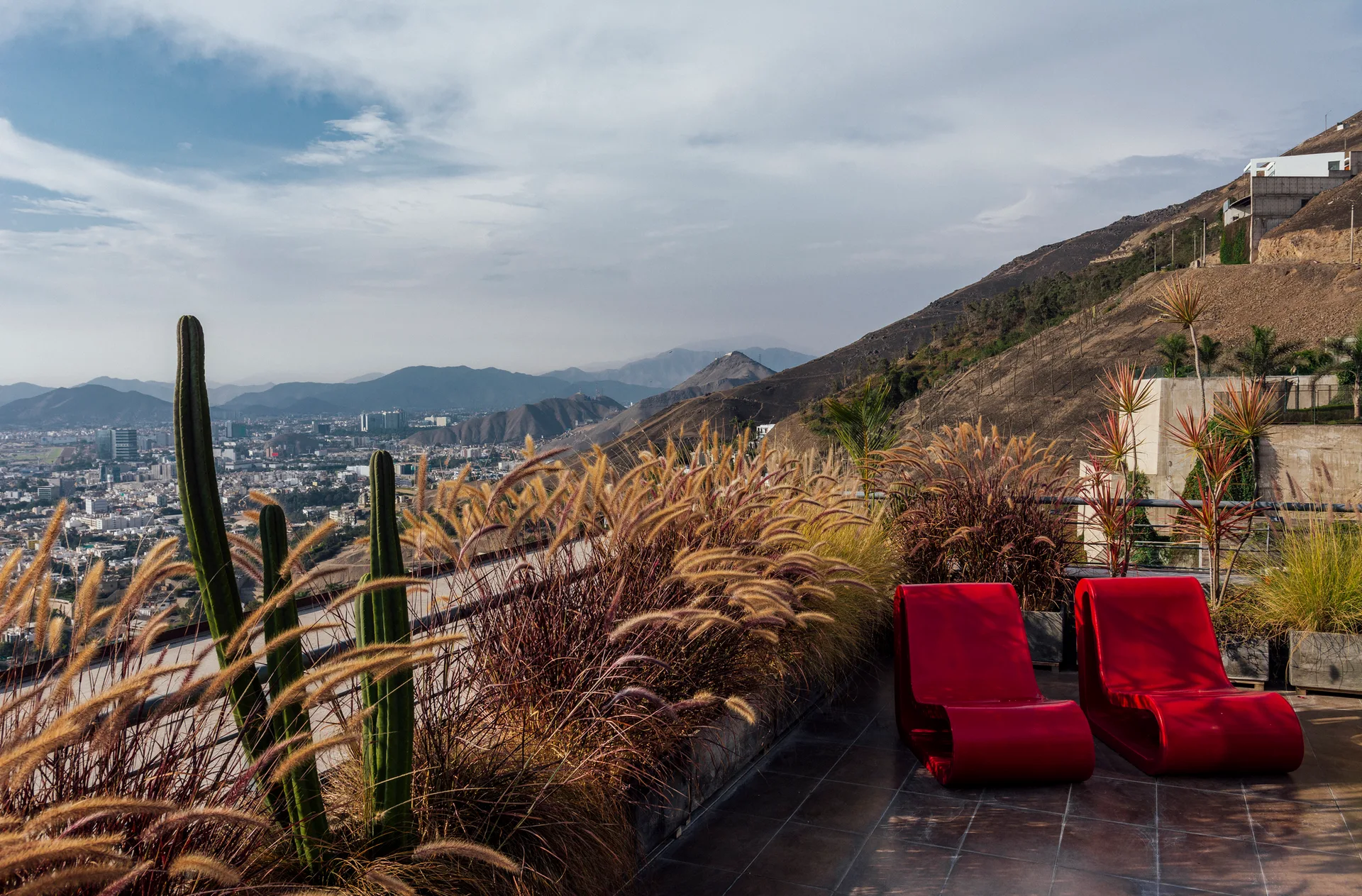 Two red curved lounge chairs sit on a tiled terrace beside planters of wild grasses, overlooking a valley city and dry mountains.