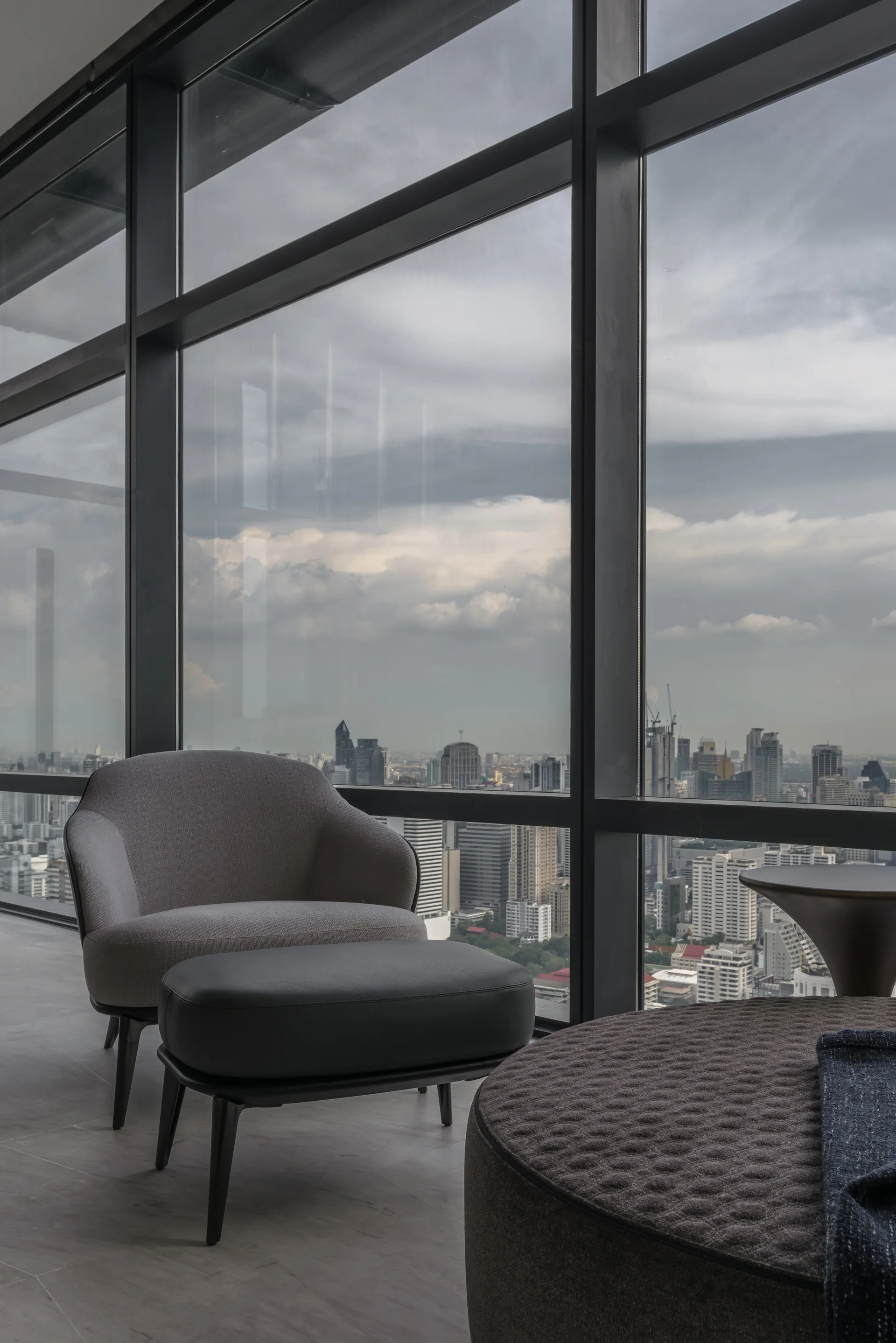 A grey fabric armchair and matching leather ottoman sit in front of a massive floor-to-ceiling window showcasing a city skyline.