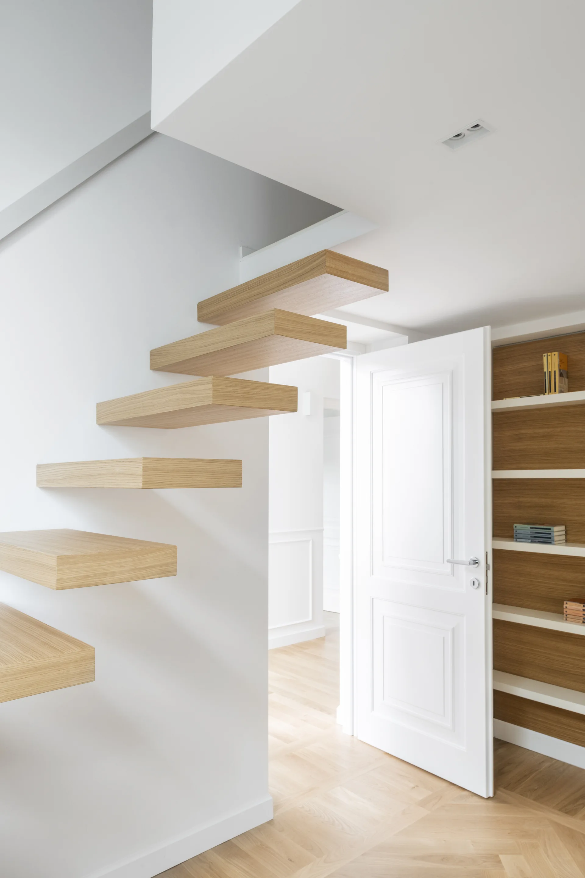 A modern hallway features cantilevered light oak stair treads on a white wall, an open white paneled door, and herringbone wood floors.