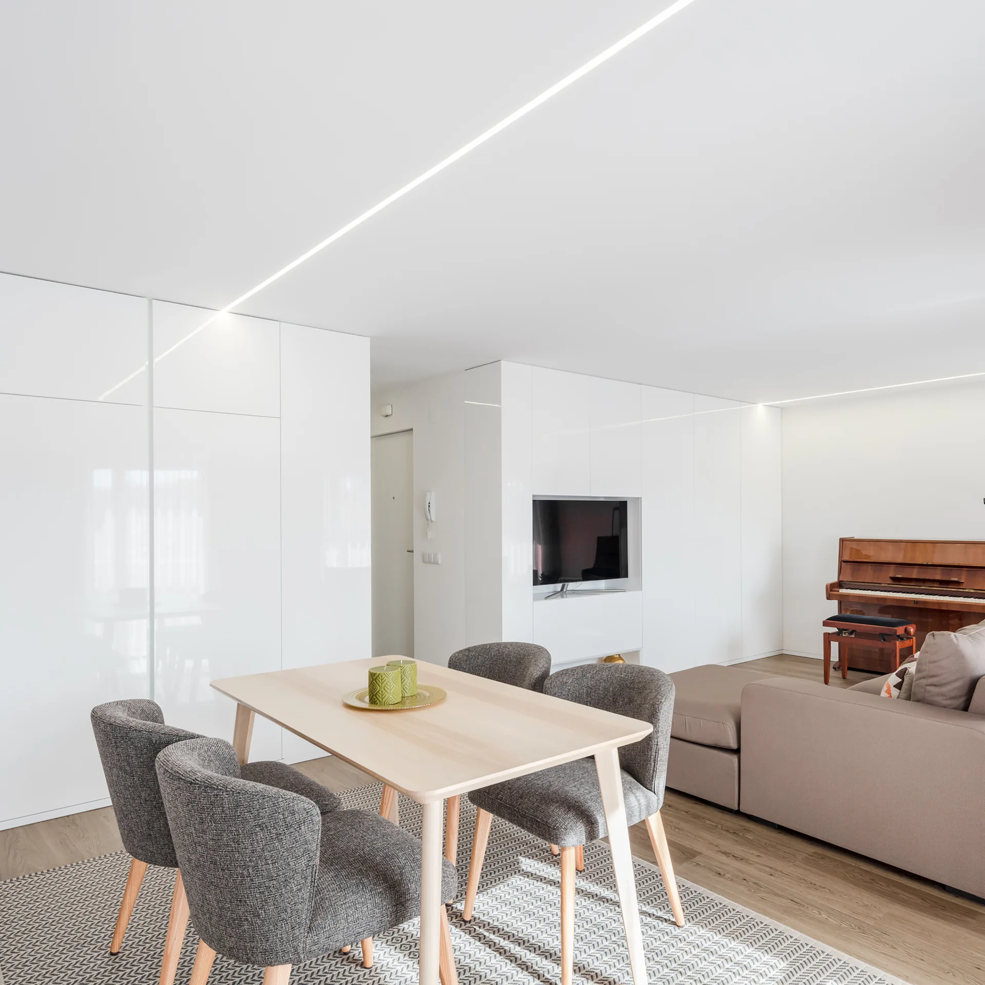 A modern open-plan living room with a light wood dining table, grey chairs, white built-in cabinets, a sofa, and an upright piano.