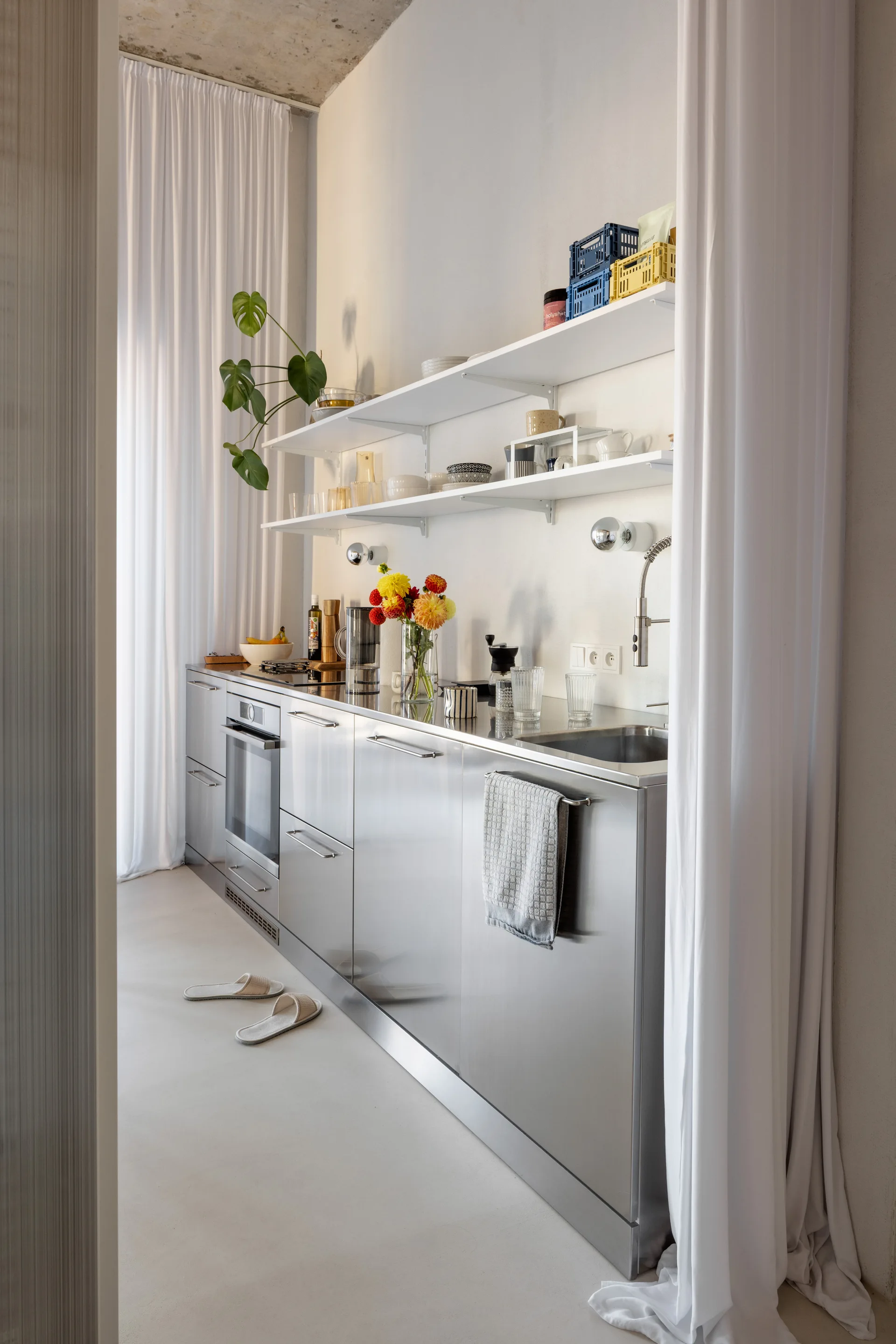 A minimalist kitchen features brushed stainless steel lower cabinets, white open shelves, and floor-to-ceiling white curtains against a concrete ceiling.