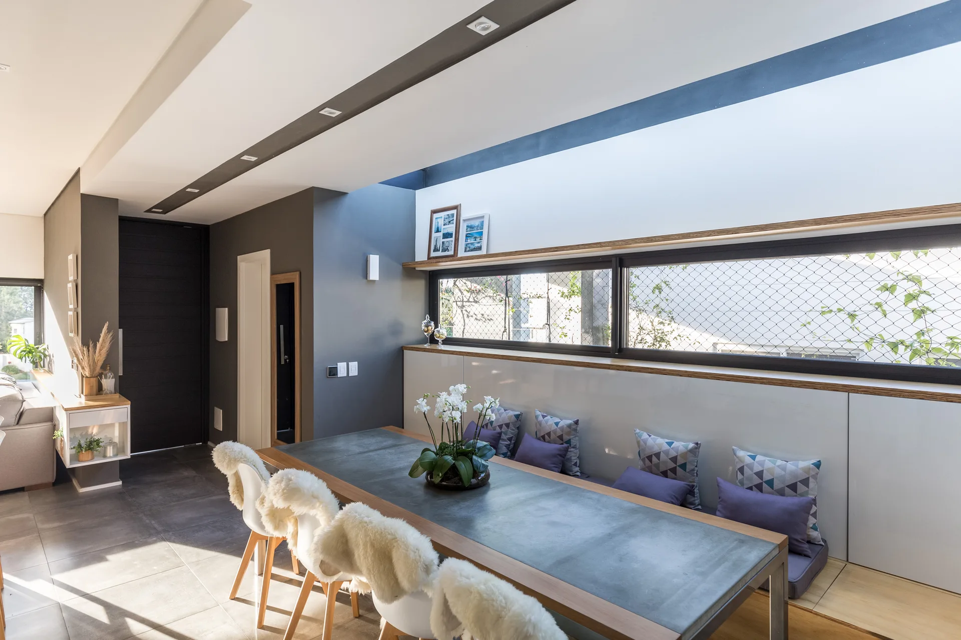 A contemporary dining room features a metal-topped dining table, white chairs with faux-fur covers, and a built-in bench beneath a long window.