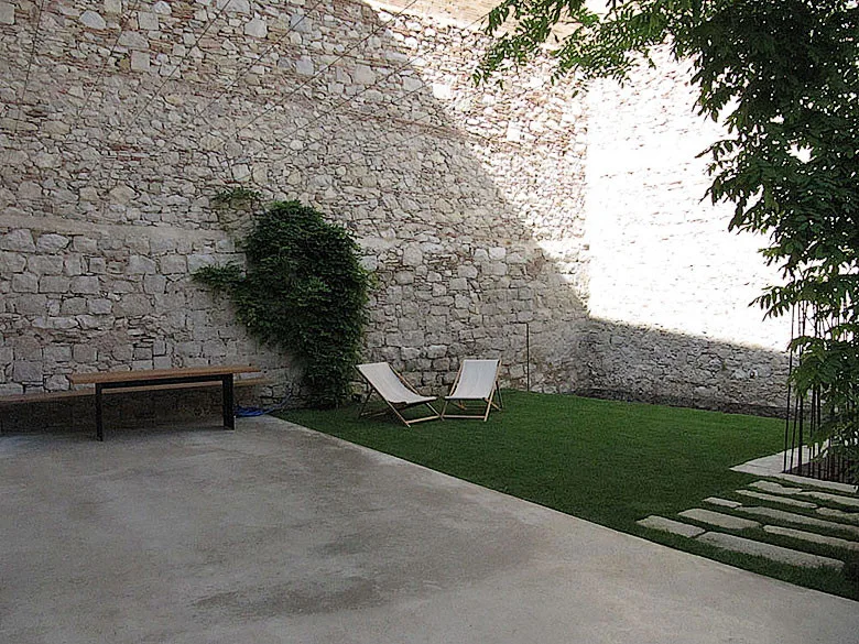 A minimalist outdoor courtyard features a tall exposed stone wall, a poured concrete patio, a small lawn with lounge chairs, and climbing green ivy.