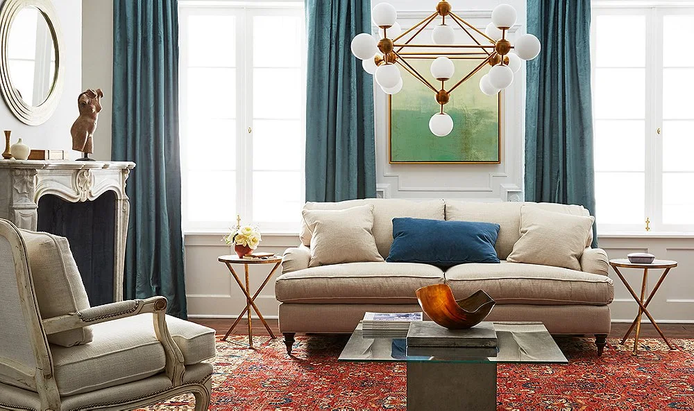 A living room featuring a beige sofa, deep teal curtains, a red patterned rug, and a modern brass chandelier hanging from the ceiling.