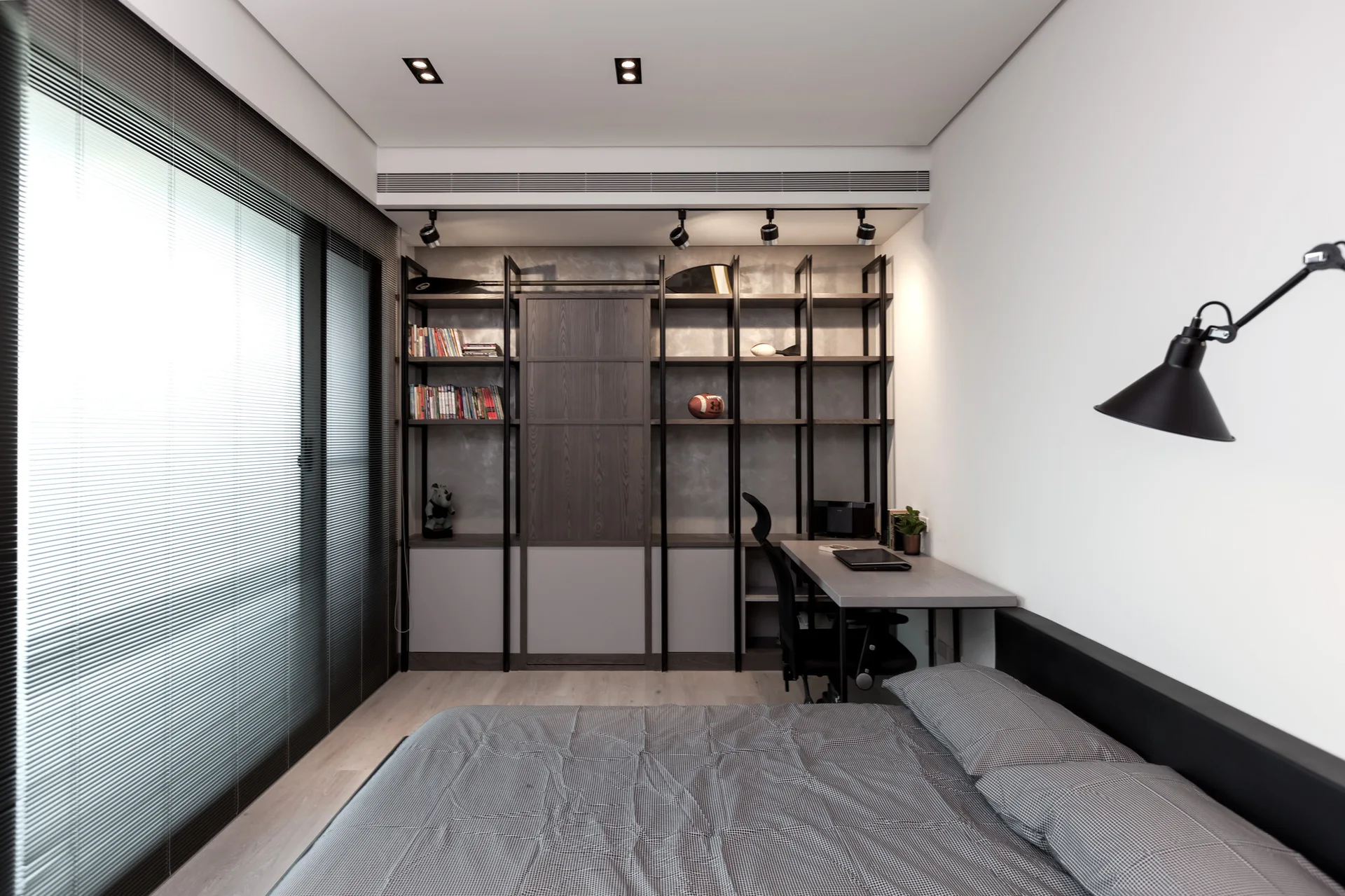 A modern bedroom featuring a bed with grey patterned bedding, a large black metal shelving unit with an integrated desk, and horizontal window blinds.