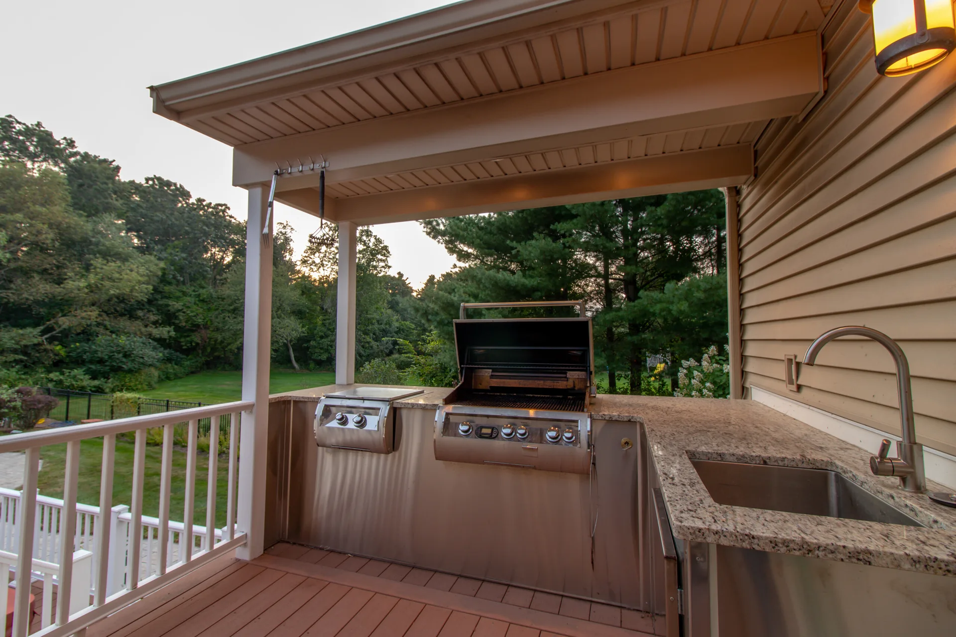 An outdoor kitchen on a covered wooden deck features a built-in stainless steel grill, a sink, and speckled stone countertops.